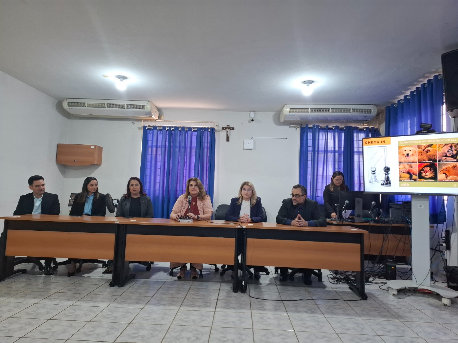 A imagem mostra uma sala de conferências com sete pessoas sentadas atrás de uma mesa longa. A mesa está dividida em duas partes, com um espaço no meio. As pessoas estão vestidas formalmente, sugerindo que pode ser uma reunião ou apresentação oficial. Ao fundo, há duas janelas com cortinas azuis e duas unidades de ar-condicionado montadas na parede. À direita, há uma tela de projeção com uma apresentação exibida. A apresentação na tela tem o título "CHECK-IN" e mostra várias imagens de animais, incluindo um cachorro, um gato e um hamster. Há também um gráfico ou diagrama na parte esquerda da tela de projeção. A sala tem piso de azulejo claro e iluminação de teto.