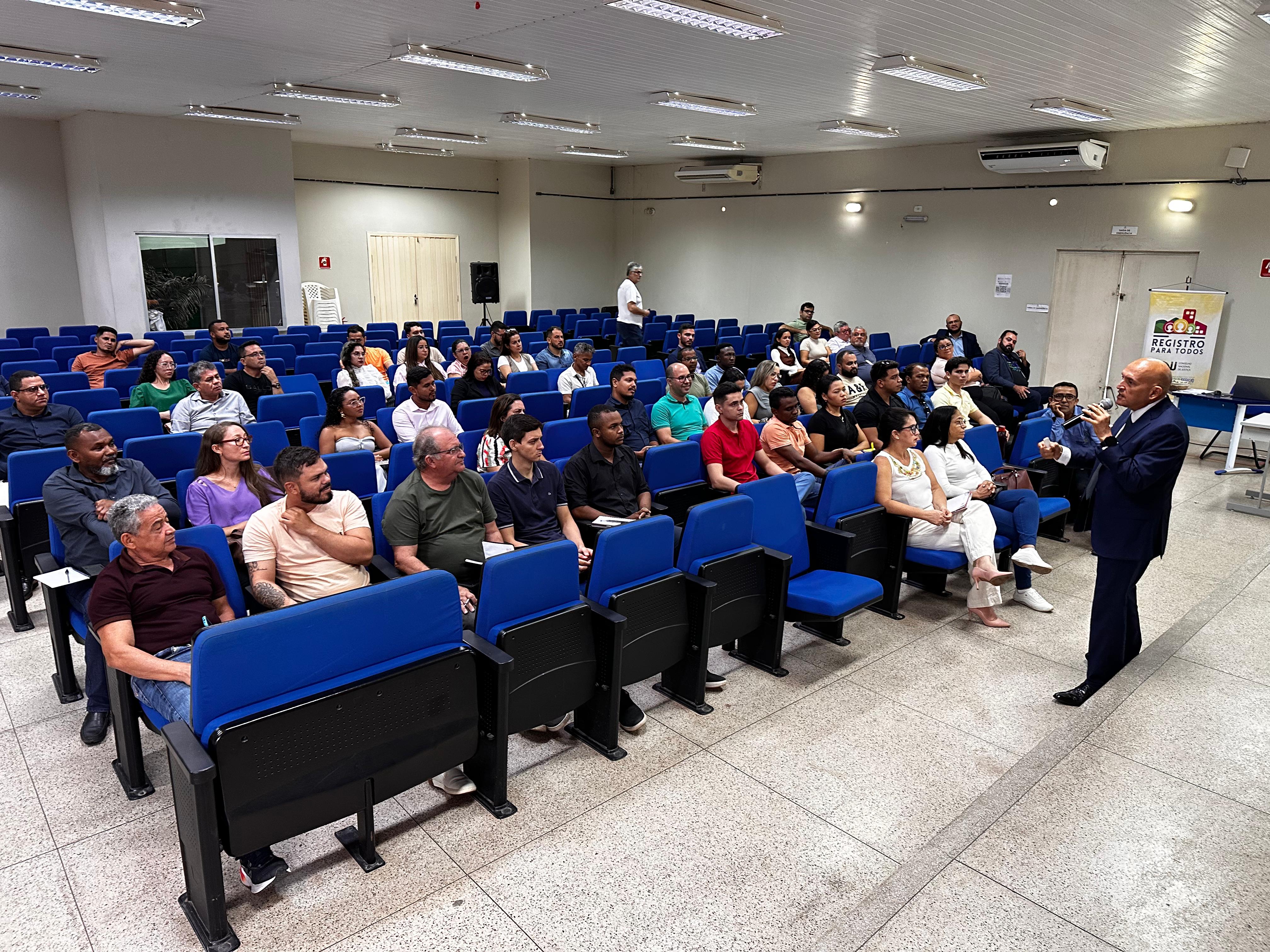 A imagem mostra uma sala de conferências com várias pessoas sentadas em cadeiras azuis, assistindo a uma apresentação. Um homem de terno está em pé na frente da sala, falando para o público. Ao fundo, há um cartaz com o texto "REGISTRO PARA TODOS" e um logotipo. A sala é bem iluminada com luzes fluorescentes no teto, e há um ar-condicionado montado na parede. As pessoas parecem estar prestando bastante atenção ao orador.