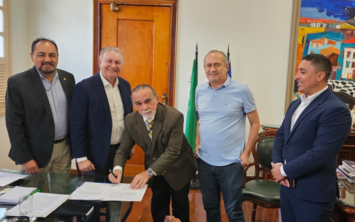 A imagem mostra um grupo de cinco homens em um ambiente de escritório. Um dos homens está assinando um documento em uma mesa de vidro, enquanto os outros quatro homens estão ao redor dele. O homem que está assinando o documento está usando terno e gravata, enquanto os outros estão vestidos com roupas formais de negócios, alguns com ternos e outros com roupas mais casuais. O escritório tem uma porta de madeira ao fundo, duas bandeiras e uma pintura colorida na parede. Há vários documentos e um copo de água sobre a mesa.