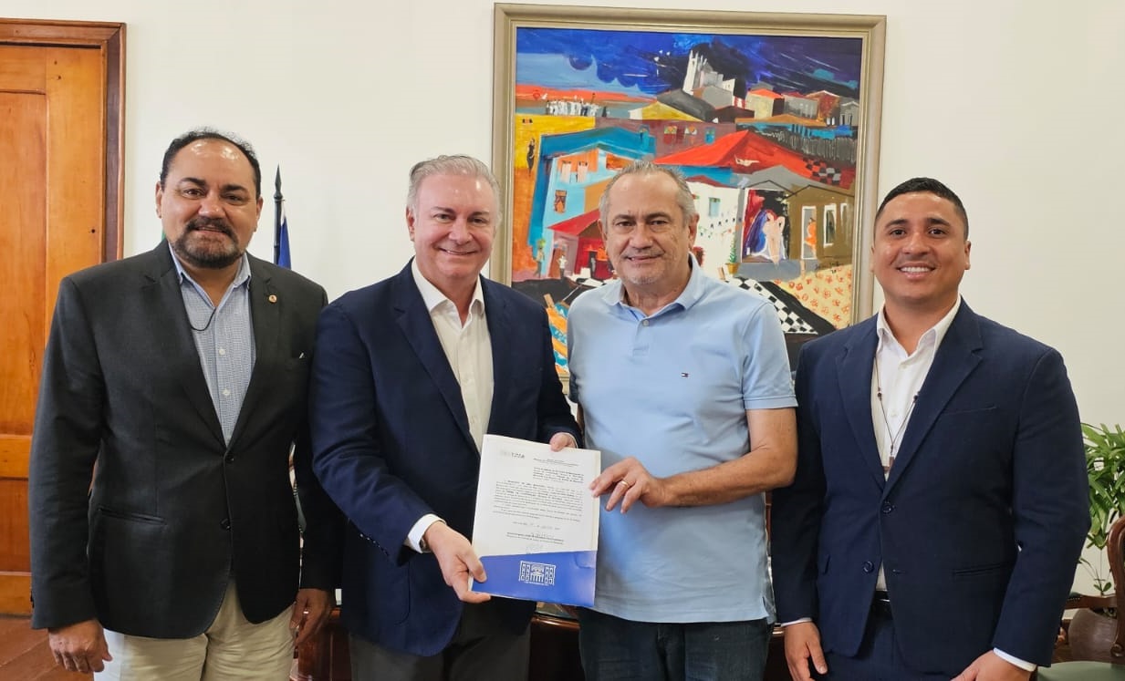 A imagem mostra quatro homens em pé, lado a lado, em um ambiente interno. Dois dos homens, que estão no centro da imagem, seguram um documento. O homem à esquerda do centro está vestindo um terno azul escuro e camisa branca, enquanto o homem à direita do centro está vestindo uma camisa polo azul claro. Os outros dois homens, um à extrema esquerda e outro à extrema direita, também estão vestidos formalmente com ternos. Ao fundo, há uma pintura colorida emoldurada na parede, que parece retratar uma cena urbana com casas e figuras humanas. O ambiente parece ser um escritório ou uma sala de reuniões, com uma porta de madeira à esquerda e uma planta à direita.