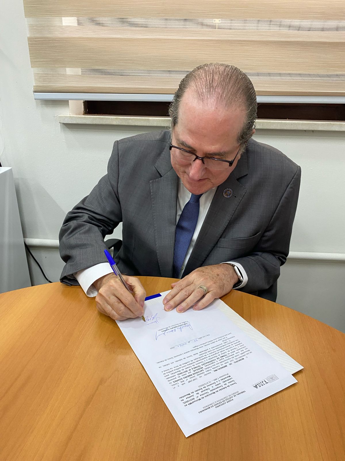 Homem vestindo terno, está sentado enquanto assina um documento