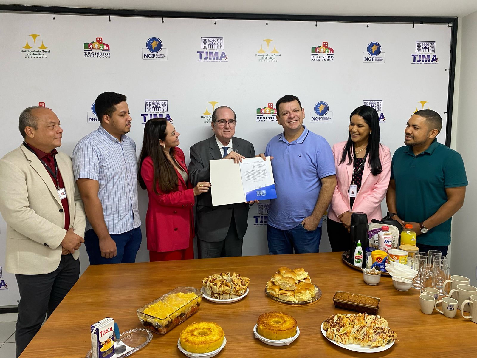 A imagem mostra um grupo de sete pessoas em pé diante de uma parede decorada com diversos logotipos, incluindo "Corregedoria Geral da Justiça," "Registro Para Todos," "NGF" e "TJMA." O grupo parece estar participando de um evento formal ou cerimônia. Uma pessoa no centro está segurando um documento, enquanto outra aponta para ele, sugerindo que o documento seja de destaque no momento.

Na frente deles, há uma mesa com vários itens alimentícios, como pães, bolos, salgados, uma caixa de suco Tirol e uma bandeja com copos e bebidas. É uma cena que transmite um encontro celebrativo ou institucional, possivelmente relacionado às organizações representadas pelos logotipos no fundo.