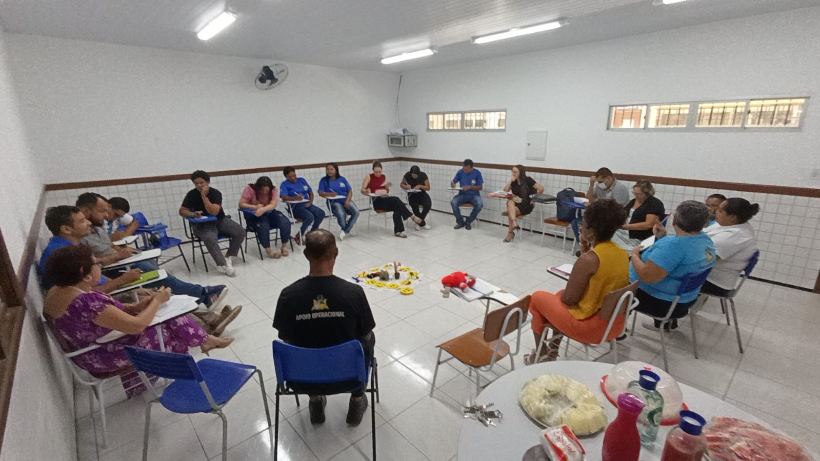 A imagem mostra um grupo de pessoas reunidas em círculo, em um ambiente que parece ser uma sala de aula ou um espaço para treinamentos e encontros. As paredes são brancas, parcialmente cobertas por azulejos na parte inferior, e há janelas altas que iluminam o local. No centro do círculo, objetos diversos estão espalhados no chão, incluindo brinquedos e materiais de atividades. Algumas pessoas estão fazendo anotações em cadernos ou pranchetas, sugerindo que participam de uma discussão ou aprendizado. Há também uma mesa ao fundo com garrafas e pratos cobertos, indicando que há alimentos e bebidas disponíveis. O ambiente é iluminado por luzes fluorescentes no teto.