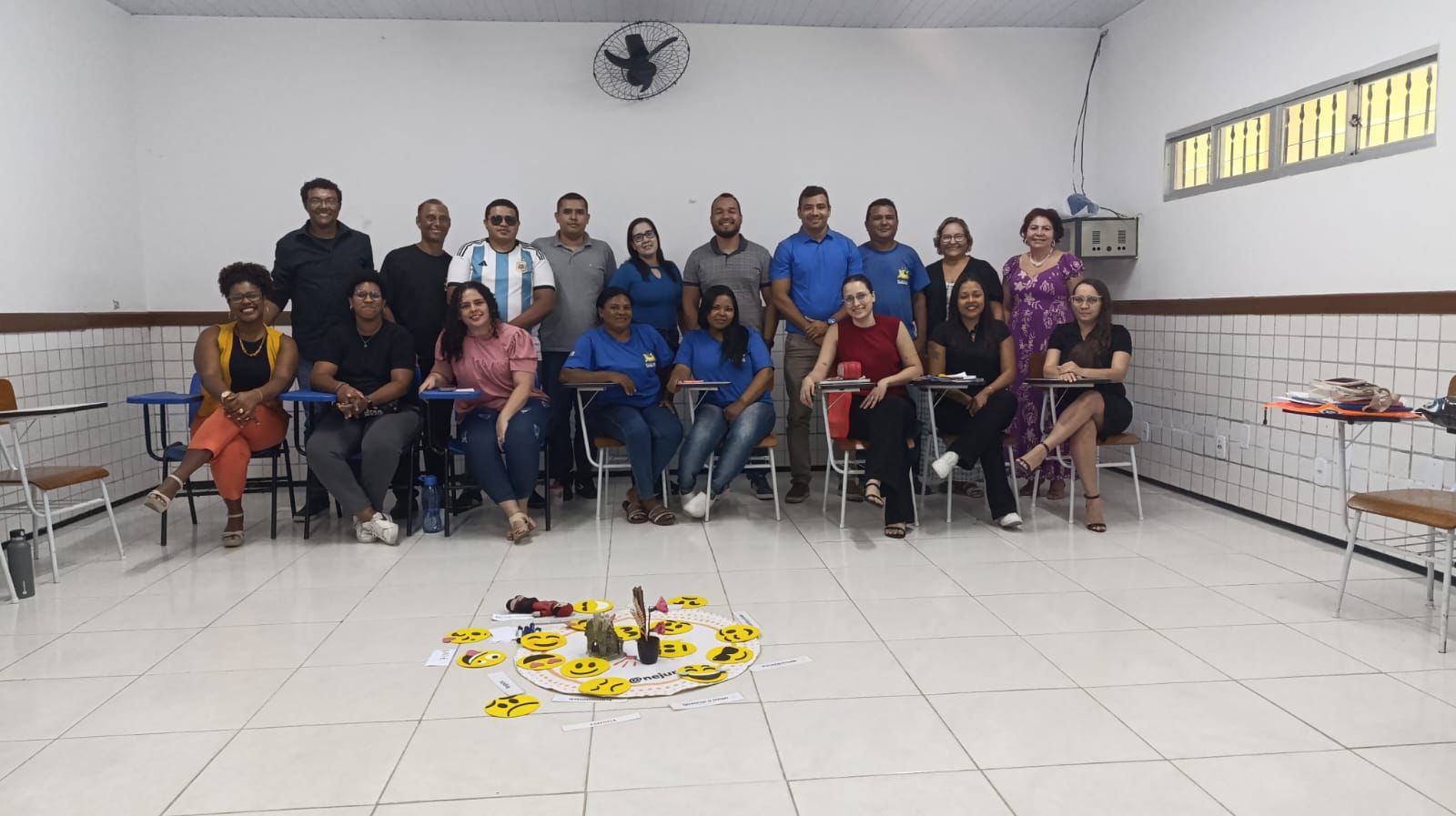 A imagem mostra um grupo de 19 pessoas reunidas em uma sala de aula, tirando uma foto em conjunto. Algumas estão sentadas em cadeiras escolares, enquanto outras estão em pé atrás delas. No ambiente, as paredes são brancas, há um ventilador de teto e janelas com grades, proporcionando uma sensação de simplicidade típica de salas de aula.  No chão, em frente ao grupo, existe um círculo feito de papéis com emojis amarelos espalhados ao redor, além de objetos como canetas e outros materiais no centro. O grupo parece estar participando de uma atividade colaborativa ou dinâmica, com um clima descontraído e interativo.