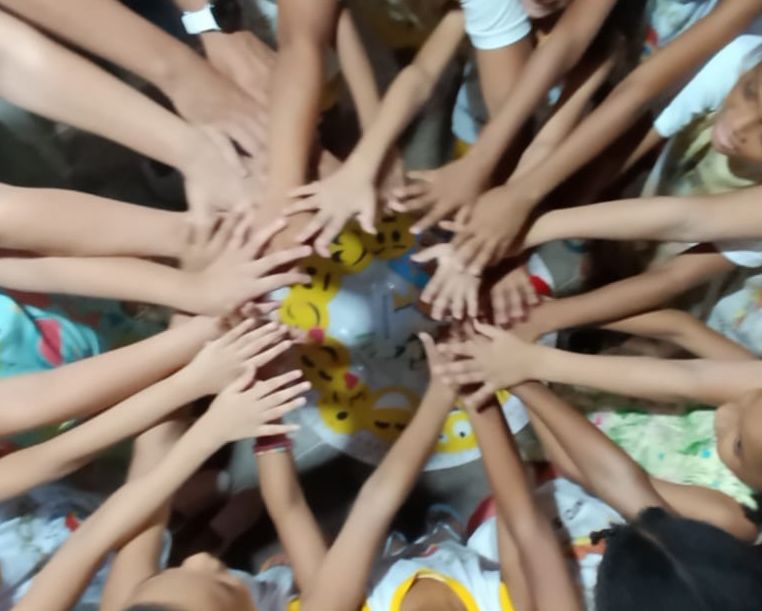 imagem fotográfica de várias mãos de crianças em circulo. no chao, um tapete com acessórios