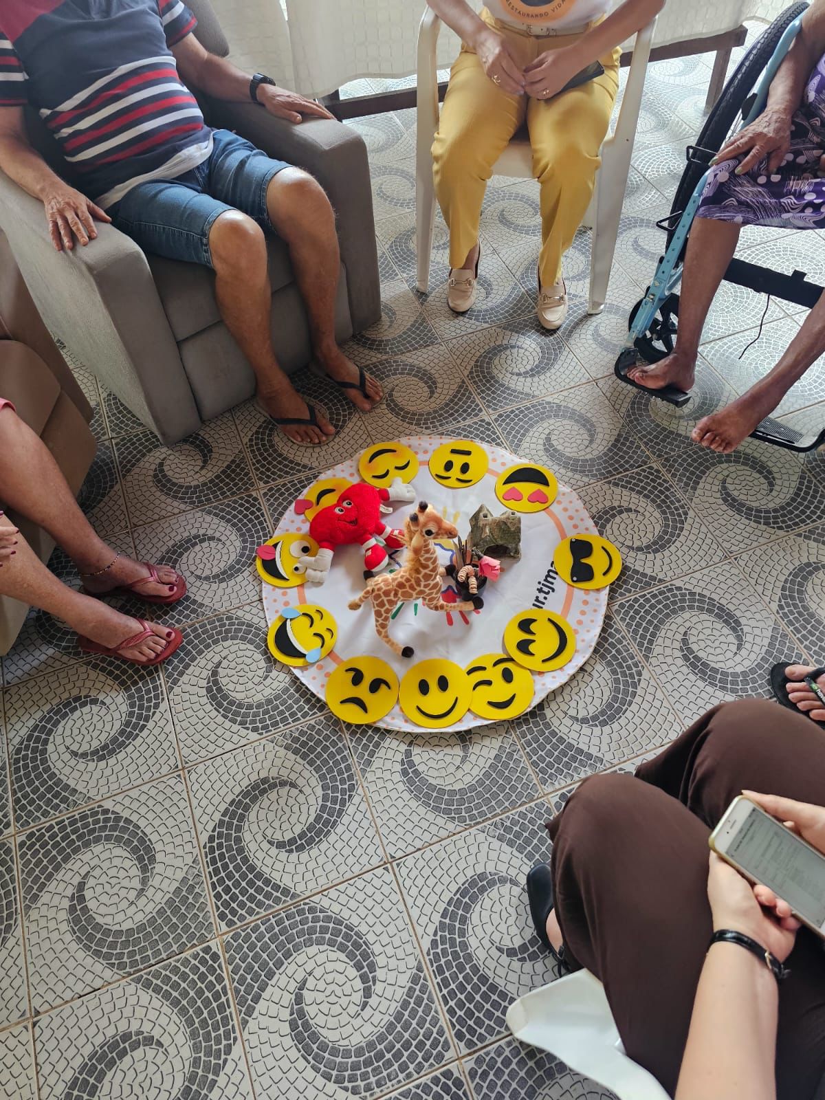 A imagem mostra um pequeno grupo de seis pessoas reunidas em círculo em um ambiente descontraído, como uma sala de convivência. Elas estão sentadas em cadeiras e sofás, envolvidas em uma atividade, possivelmente relacionada à comunicação ou expressão emocional. No centro do círculo, há um tecido branco no chão, sobre o qual estão colocados rostos de emojis que expressam várias emoções, como alegria, tristeza e riso.  Além disso, há alguns bichos de pelúcia, incluindo uma girafa, uma tartaruga verde e outro personagem vermelho, dispostos de maneira a chamar a atenção. O piso da sala tem um padrão de azulejos com desenhos ondulados em tons de cinza e branco.