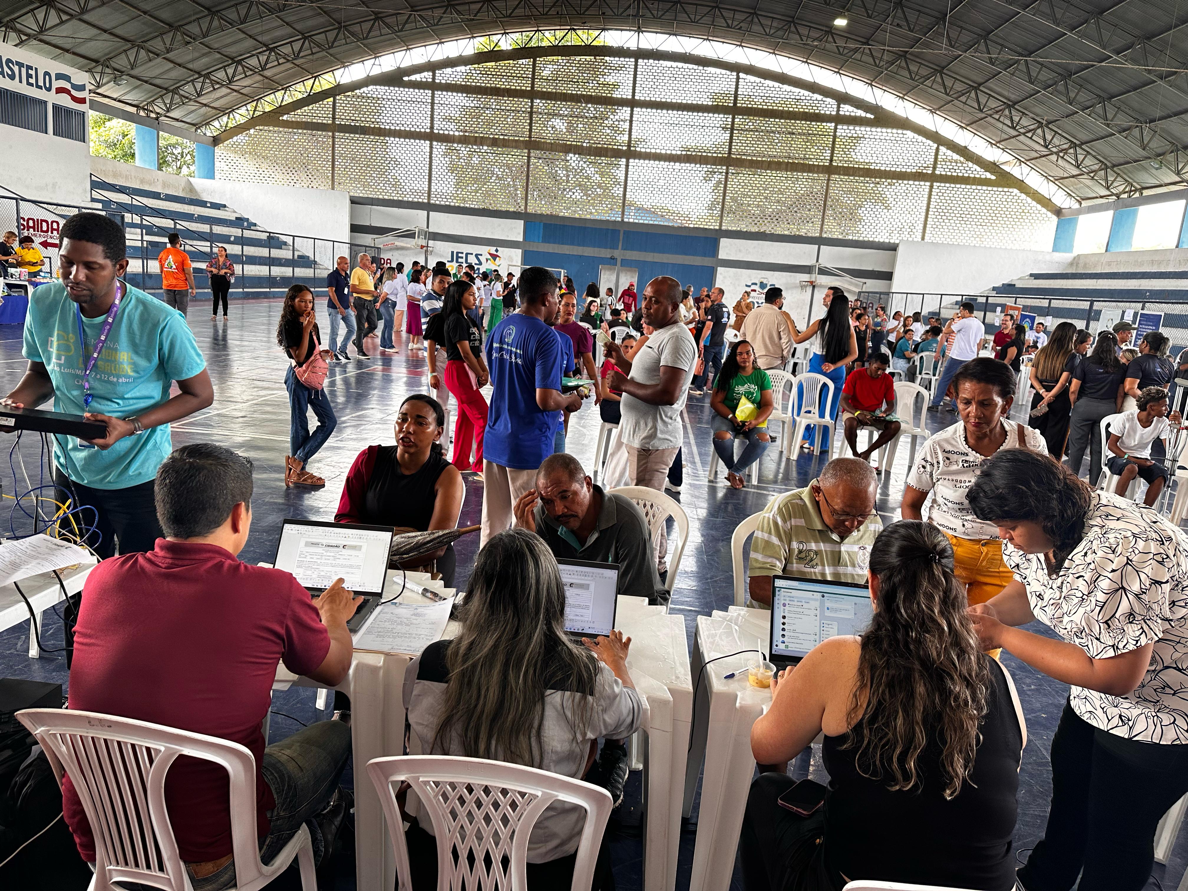 A imagem mostra um espaço amplo, como um ginásio ou centro comunitário, onde está acontecendo um evento público. Há várias mesas organizadas, com pessoas sentadas trabalhando em laptops, aparentemente ajudando outros participantes. Muitas outras pessoas estão de pé ou sentadas em cadeiras, esperando ou conversando. O ambiente parece estruturado e organizado, com um número significativo de participantes.O evento parece estar focado em fornecer serviços ou informações à comunidade, dado o uso de computadores e papéis. A atmosfera sugere colaboração e interação social.