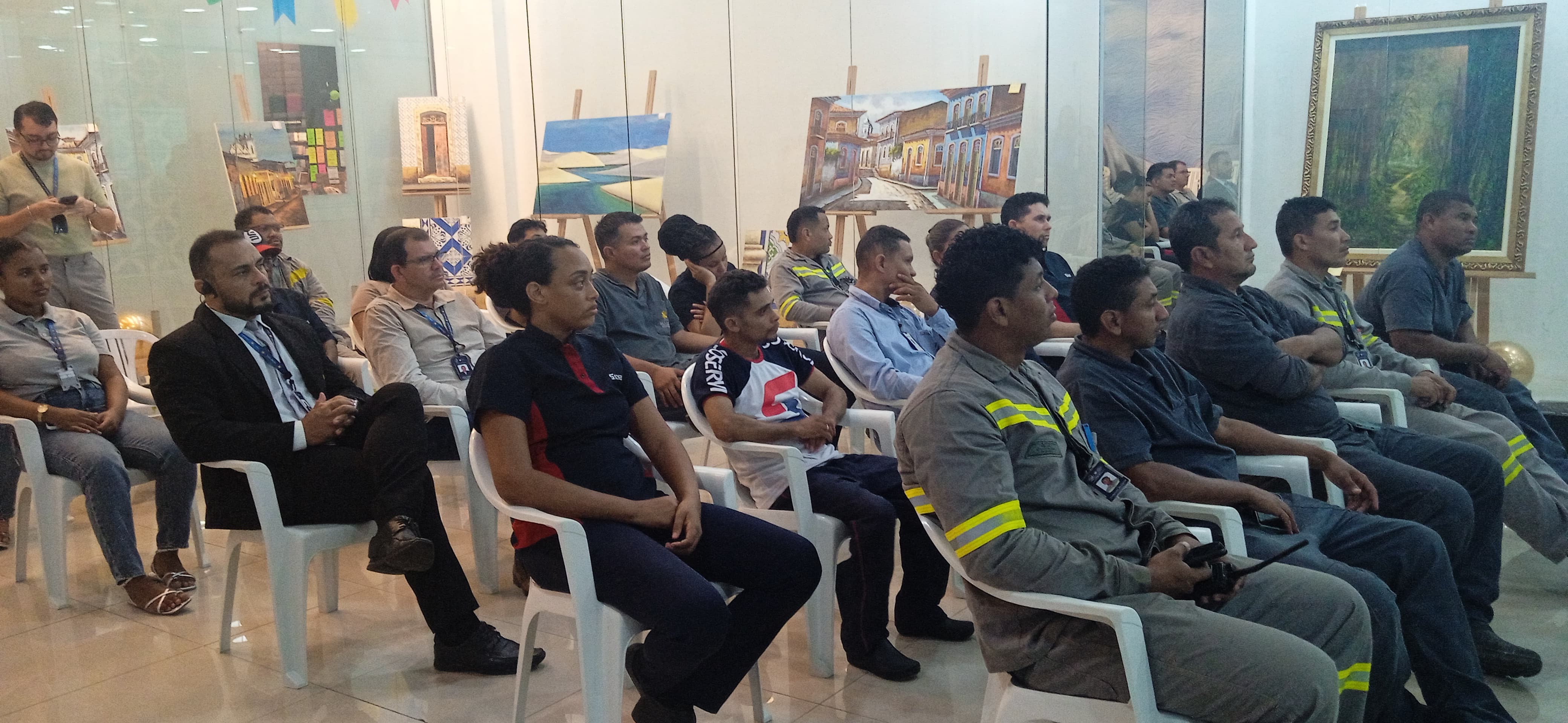 Pessoas sentadas em cadeiras plásticas brancas assistem a uma apresentação em ambiente interno. O público é formado por homens e mulheres, muitos com uniformes de manutenção. Ao fundo, há quadros com pinturas expostos em cavaletes e decoração temática.