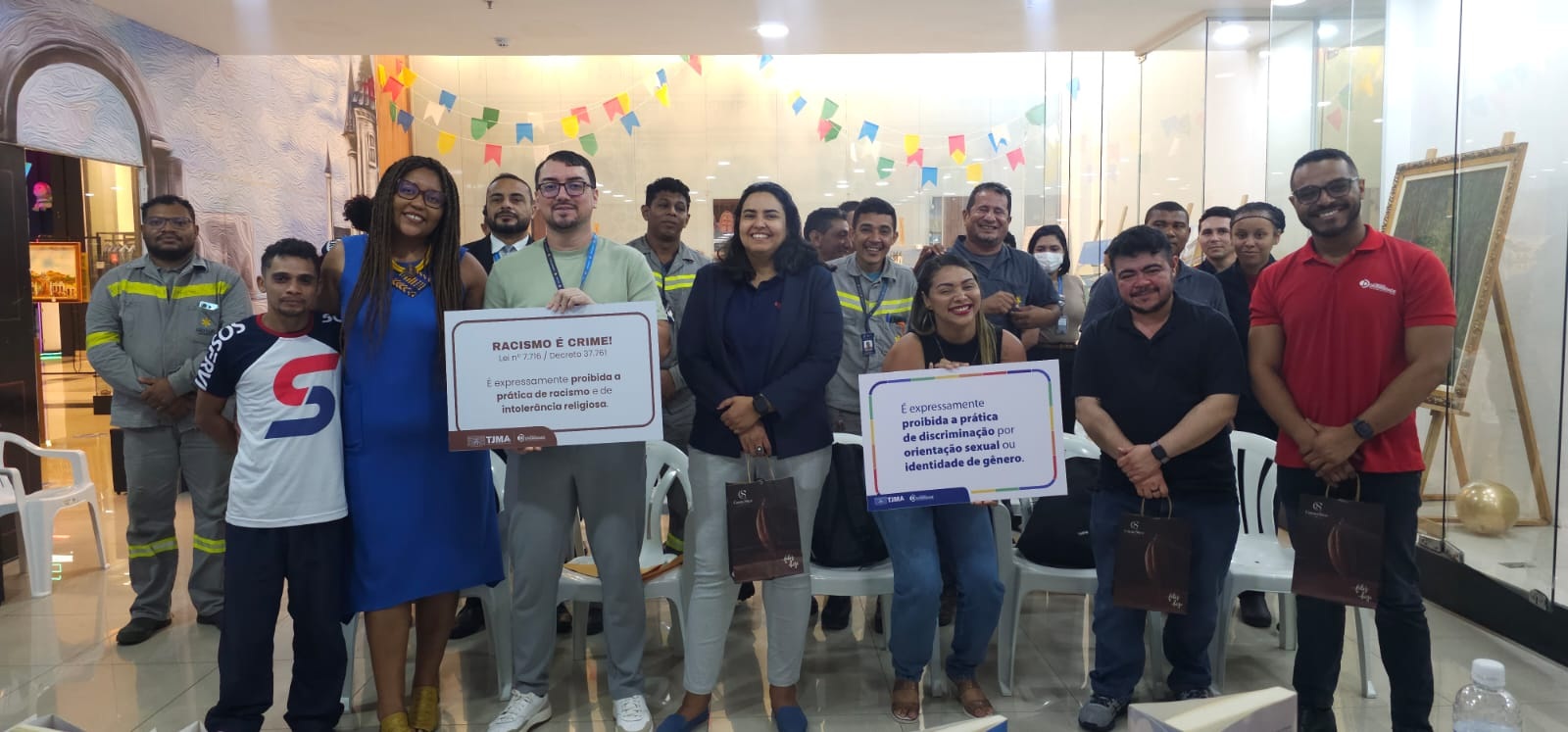 Grupo de aproximadamente 20 pessoas posa sorridente para a foto em um espaço interno, com decoração de bandeirinhas coloridas ao fundo. No centro, duas pessoas seguram cartazes com mensagens contra a discriminação: um deles diz ¿Racismo é crime!¿ e o outro, ¿É expressamente proibida a prática de discriminação por orientação sexual ou identidade de gênero¿. À frente, alguns participantes seguram sacolas. Ao fundo, há cadeiras brancas vazias e uma parede de vidro. O ambiente é iluminado e aparenta ser uma área de eventos de um shopping.