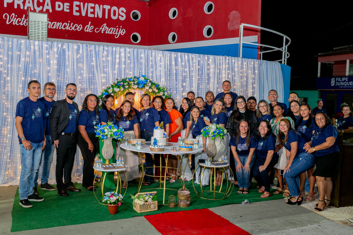 Foto horizontal, colorida, de homens e mulheres vestidos de blusa e calça azuis, em volta de mesa decorada nas cores azul, verde e dourado, com bolo nupcial. Ao fundo, cortina branca com luzes.