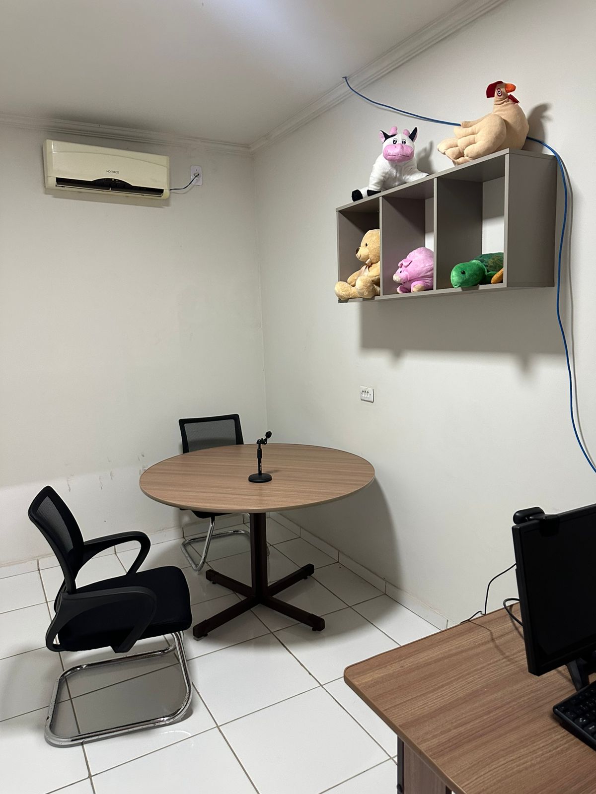 Foto vertical, colorida, mostra uma sala simples e funcional, possivelmente um escritório ou sala de reunião pequena. No centro, há uma mesa redonda de madeira clara com base em formato de X, sobre a qual repousa um microfone preto. Duas cadeiras de escritório pretas com tela respirável, uma com rodinhas e outra com base cromada, estão posicionadas ao redor da mesa.

Na parede acima da mesa, um ar-condicionado branco está instalado no canto superior esquerdo. Mais à direita, uma prateleira cinza dividida em nichos exibe vários bichos de pelúcia: uma galinha, um porco, uma tartaruga e um urso. Um cabo azul desce da parte superior da parede, próximo à prateleira. No canto inferior direito da imagem, aparece uma parte de uma mesa de madeira escura com um monitor de computador preto e um teclado. O piso da sala é revestido com ladrilhos brancos grandes e brilhantes, e as paredes são brancas