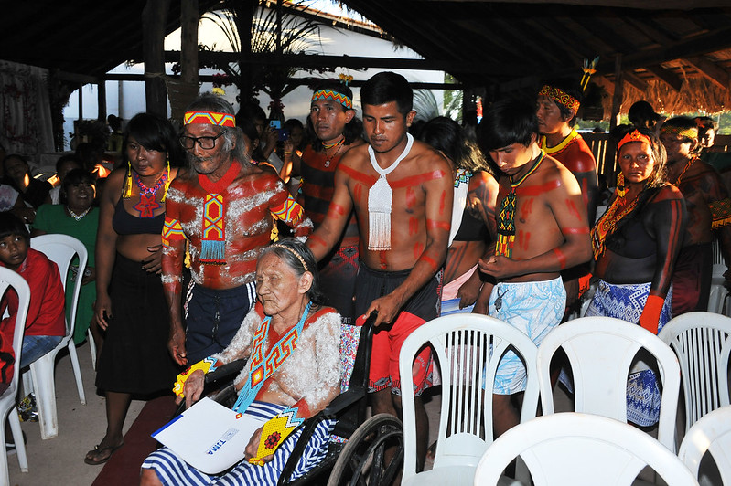 Casal de indígenas idosos, com mulher com documento nas mãos, sentados, em cadeiras brancas, junto a outros indígenas de pé, em barracão de palha