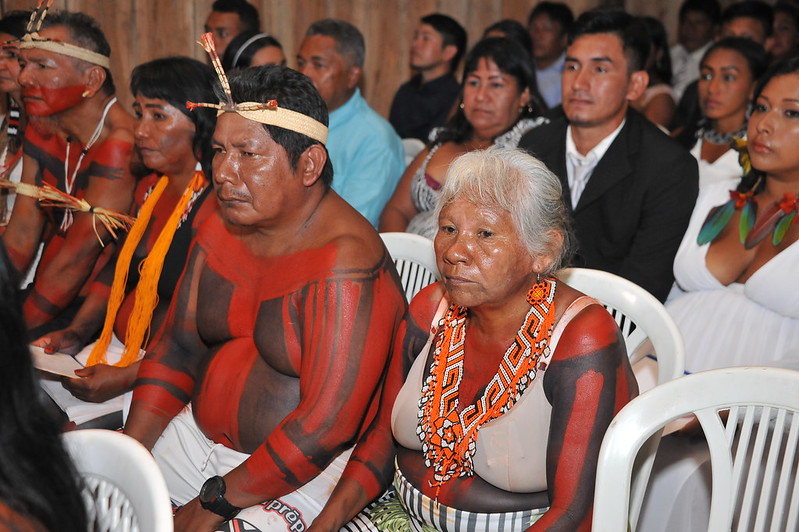 Casal de indígenas idosos sentados em cadeiras brancas em barracão de palha, junto a outros indígenas