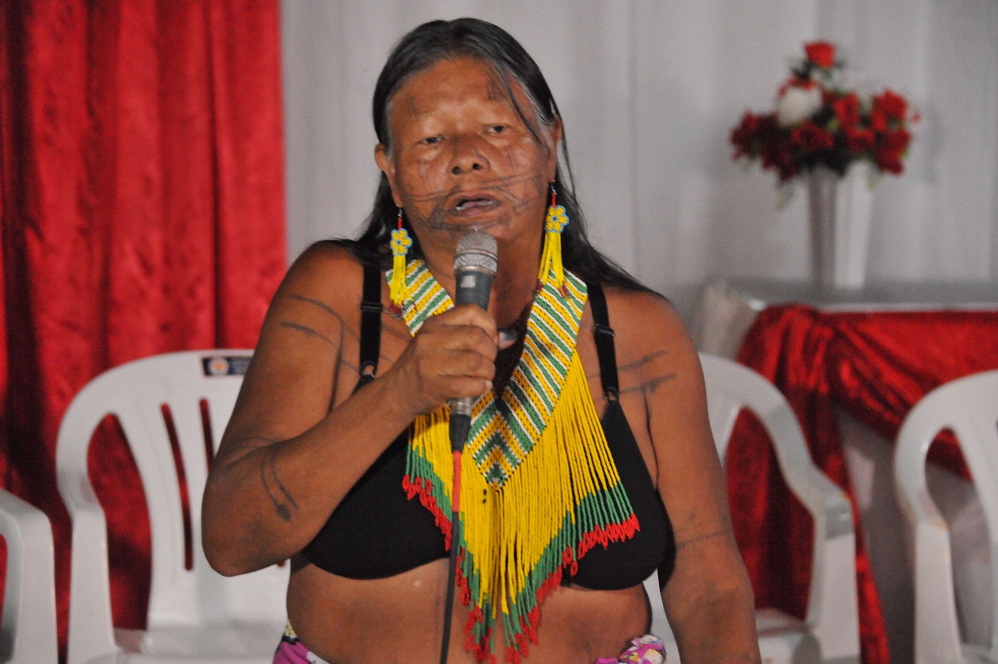 Mulher indígena idosa, de sutiã preto e colar amarelo, laranja e verde,segura microfone em salão decorado de branco e vermelho