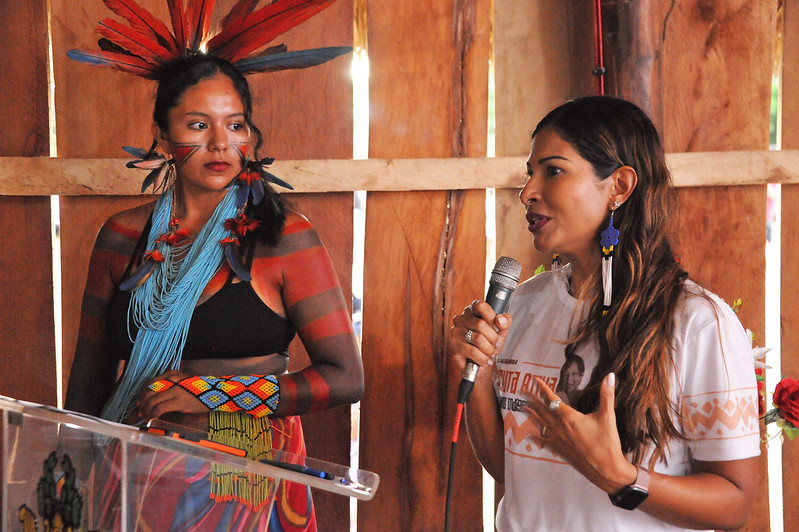 Mulher parda, de camiseta branca, de pé, segura microfone, ao lado de mulher indígena de sutiã preto e adornos coloridos, em barraca de madeira.