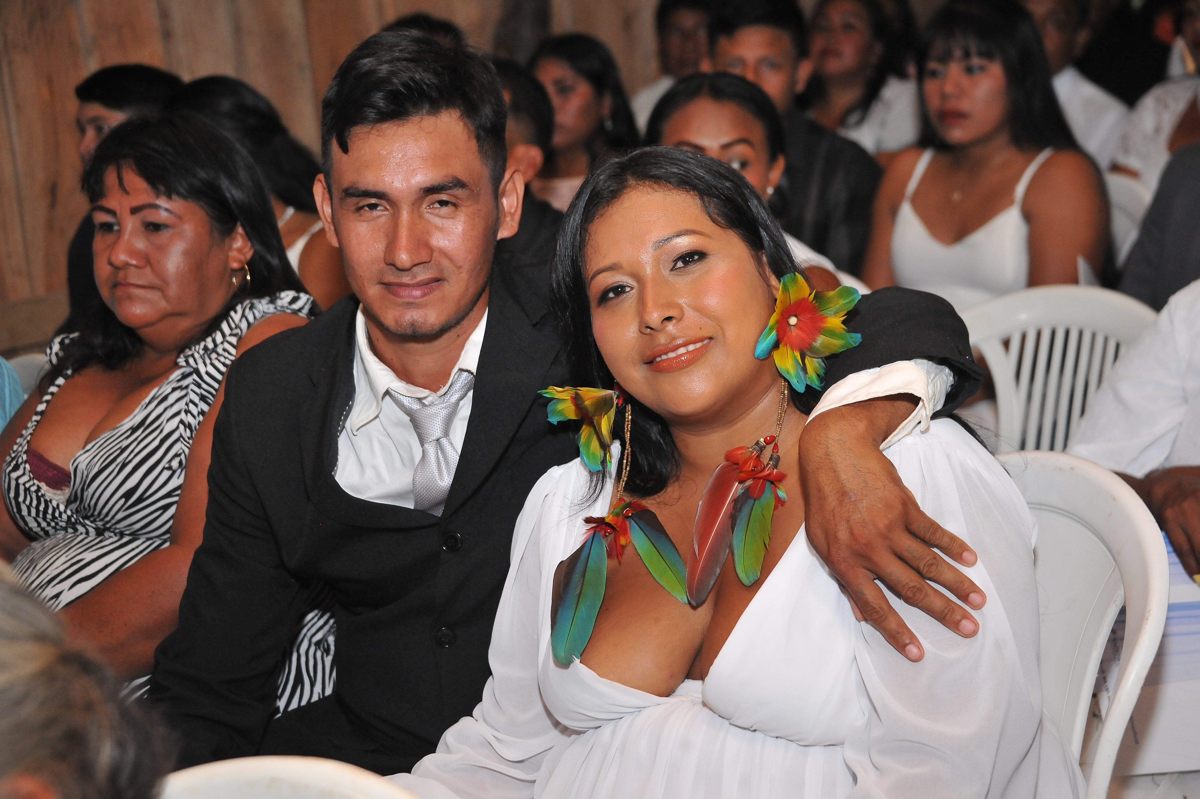 casal de jovens indígenas, ele de terno preto, ela de vestido branco e colar e brincos de penas coloridas.
