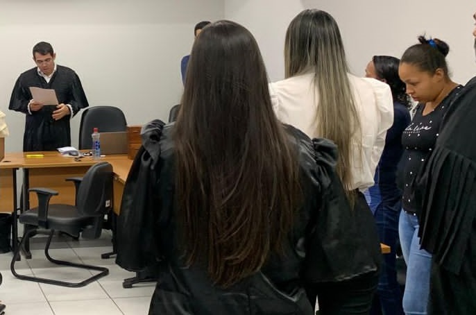 imagem na qual aparecem 4 pessoas, sendo uma de frente, um homem, e duas mulheres de costa e uma de lado, em uma sala, durante um julgamento