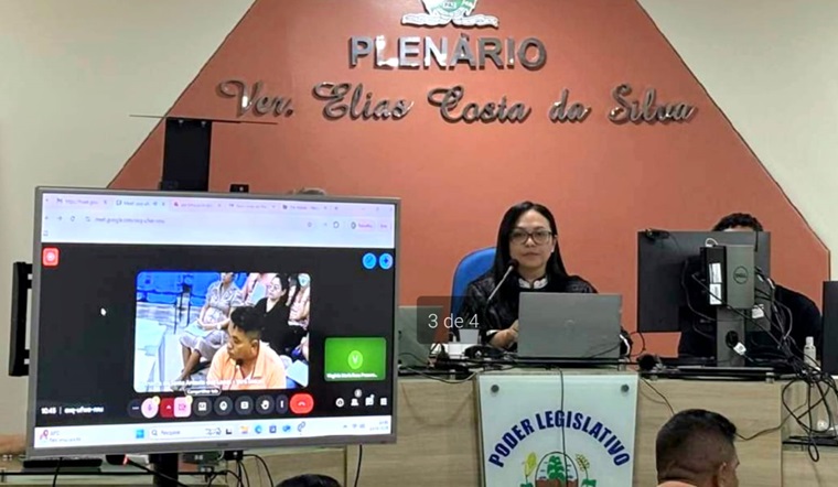 fotografia à meia distância, na qual aparecem duas pessoas sentadas atrás de uma bancada, uma mulher e um homem. ao lado deles, uma televisão e atrás um painel. eles estão em um plenário, participando de uma sessão de julgamento