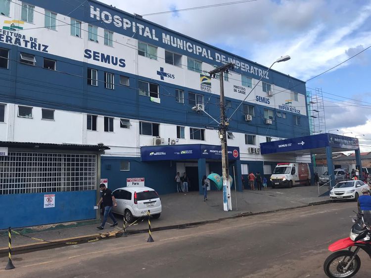 imagem na qual aparece a fachada de um hospital, com carros na frente, em uma rua movimentada