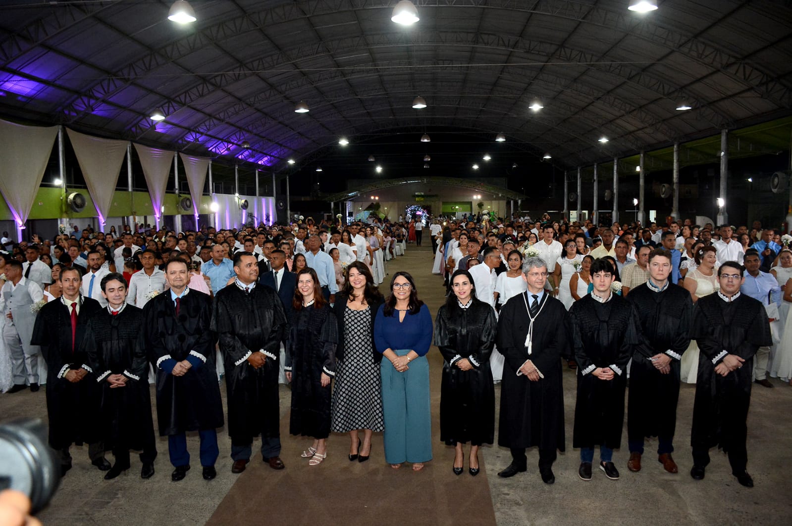 homens e mulheres de pé, vestidos de toga preta, diante de casais em galpão durante cerimônia de casamento comunitário.