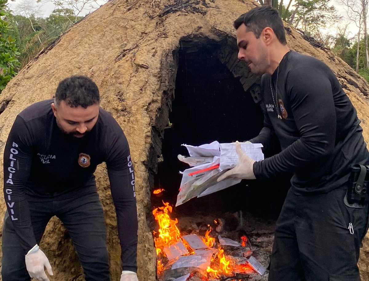 imagem na qual aparece dois homens, de preto, policiais, colocando coisas dentro de um forno em chamas, a céu aberto