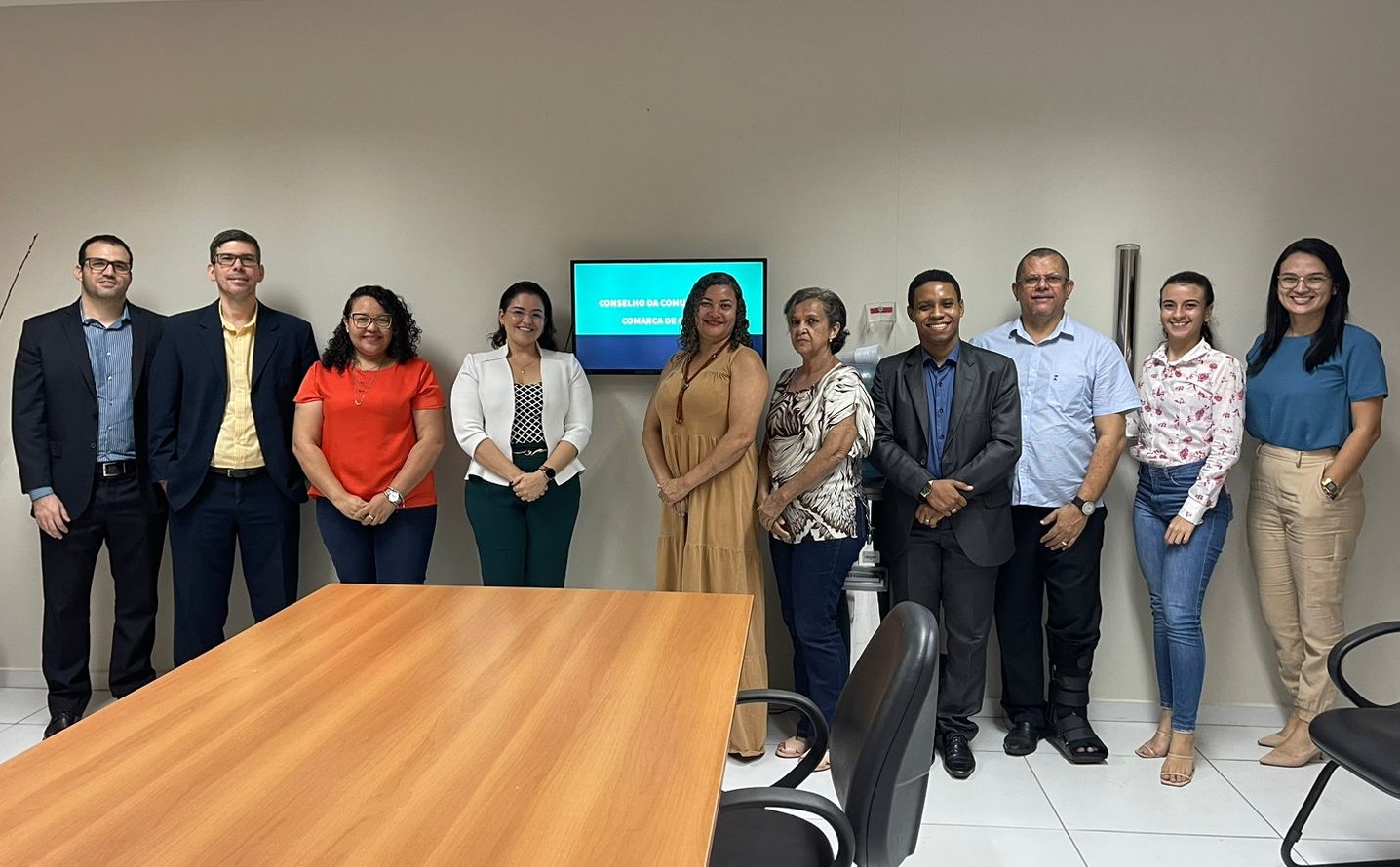 Homens e mulheres de pé, lado a lado diante de parede cinza e atrás de mesa de madeira