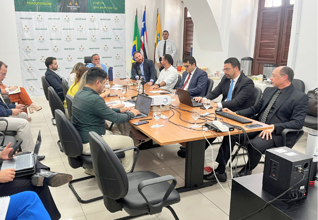Reunião institucional realizada em sala da Corregedoria Geral da Justiça do Maranhão. Ao redor de uma mesa retangular, diversos participantes ¿ entre magistrados, servidores e assessores ¿ estão reunidos com notebooks, tablets e documentos. Ao fundo, aparecem as bandeiras do Brasil, do Maranhão e do Poder Judiciário, além de um painel institucional com a identidade visual do projeto ¿Produtividade¿. Há também um projetor e equipamentos de áudio e vídeo sobre a mesa, indicando que a reunião pode estar sendo gravada ou transmitida.