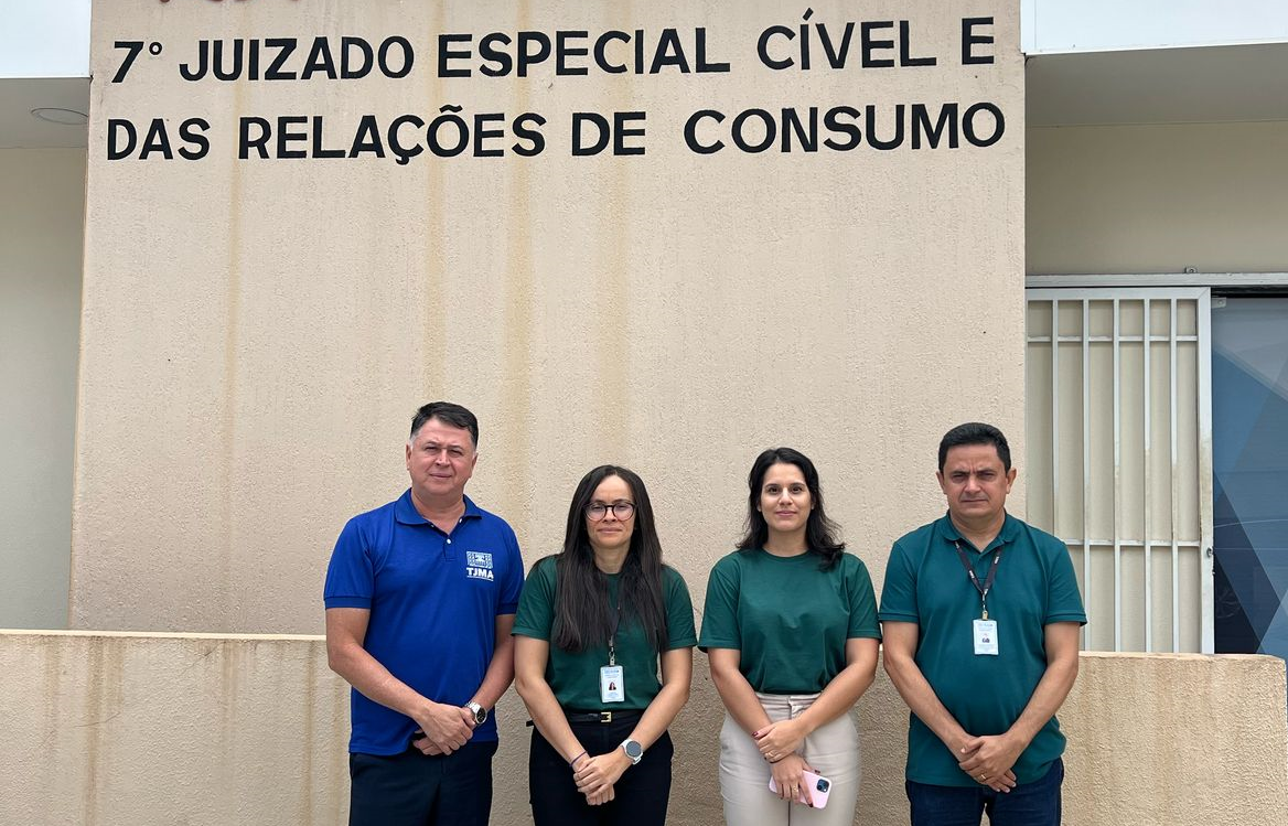 Foto horizontal, colorida, mostra quatro pessoas de pé, posando para foto, lado a lado, diante de fachada de prédio na cor bege, com o nome "7º Juizado Cível e das Relaç~çeos de Consumo". Um dos homens, o primeiro à esquerda, é branco e veste camisa azul e calça escura. Os demais - duas mulheres e um homem, vestem camisa verde.