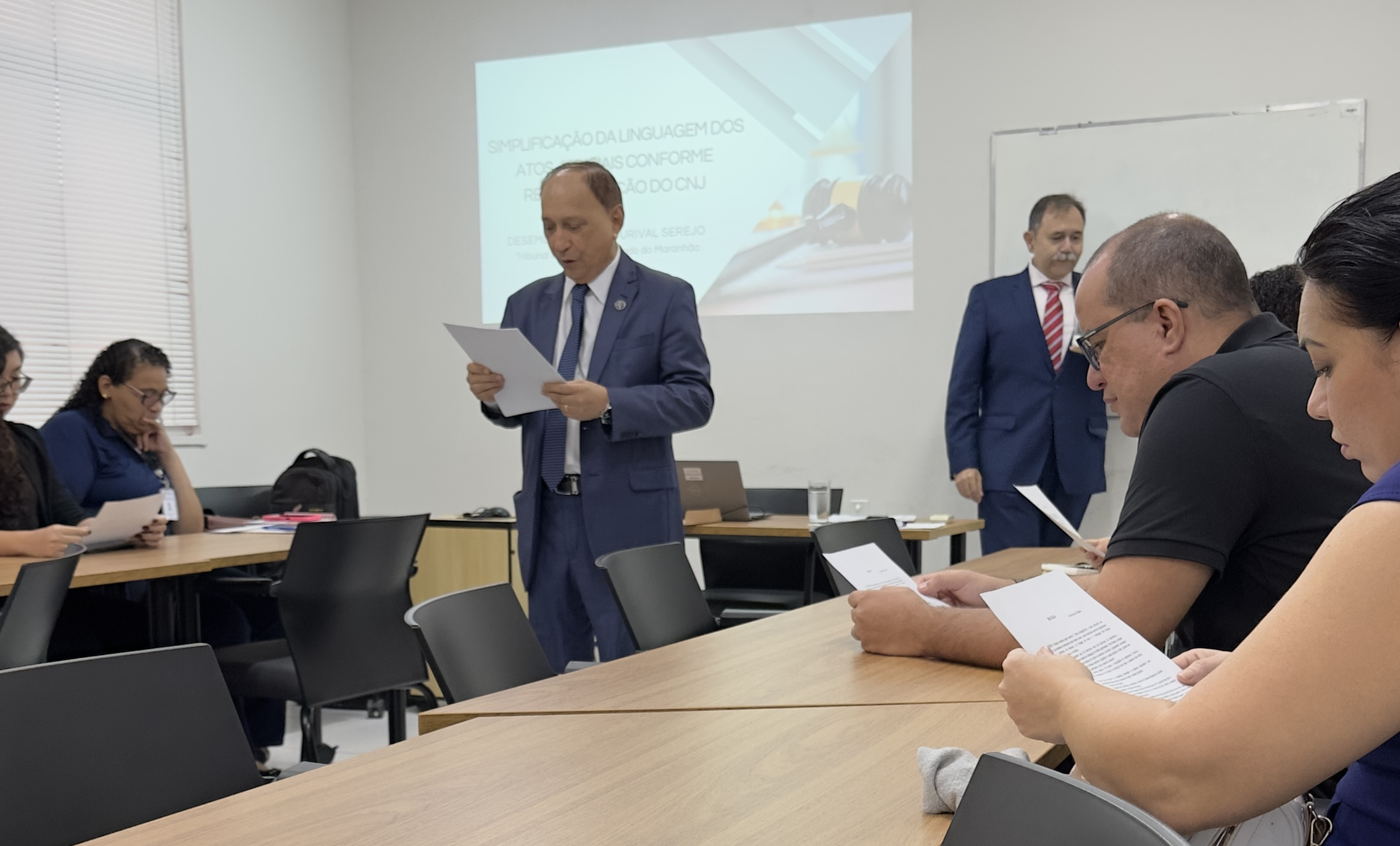 Foto horizontal, colorida, mostra um grupo de pessoas em uma sala de aula ou ambiente de reunião. Duas pessoas estão de pé na frente da sala, uma delas segurando um papel e aparentando estar falando ou apresentando algo. A outra pessoa está ligeiramente atrás e ao lado, também segurando um papel. Várias outras pessoas estão sentadas em mesas, olhando para papéis em suas mãos.

Há uma apresentação projetada na parede atrás dos palestrantes, que lê: "Simplificação da Linguagem dos Atos Processuais Conforme Recomendação do CNJ".