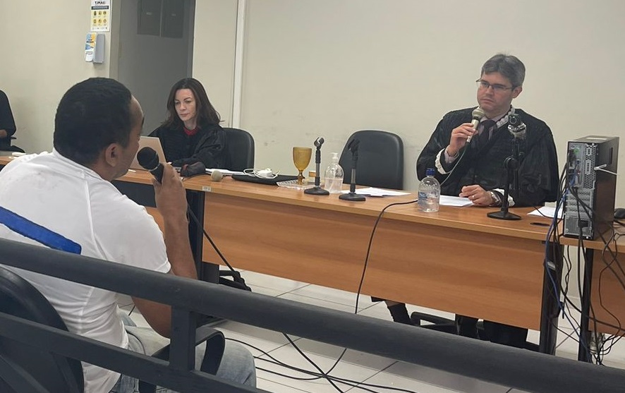 imagem na qual aparece um homem, de costas, blusa branca, e do outro lado um homem e uma mulher, atras de uma mesa, durante  uma sessão e julgamento