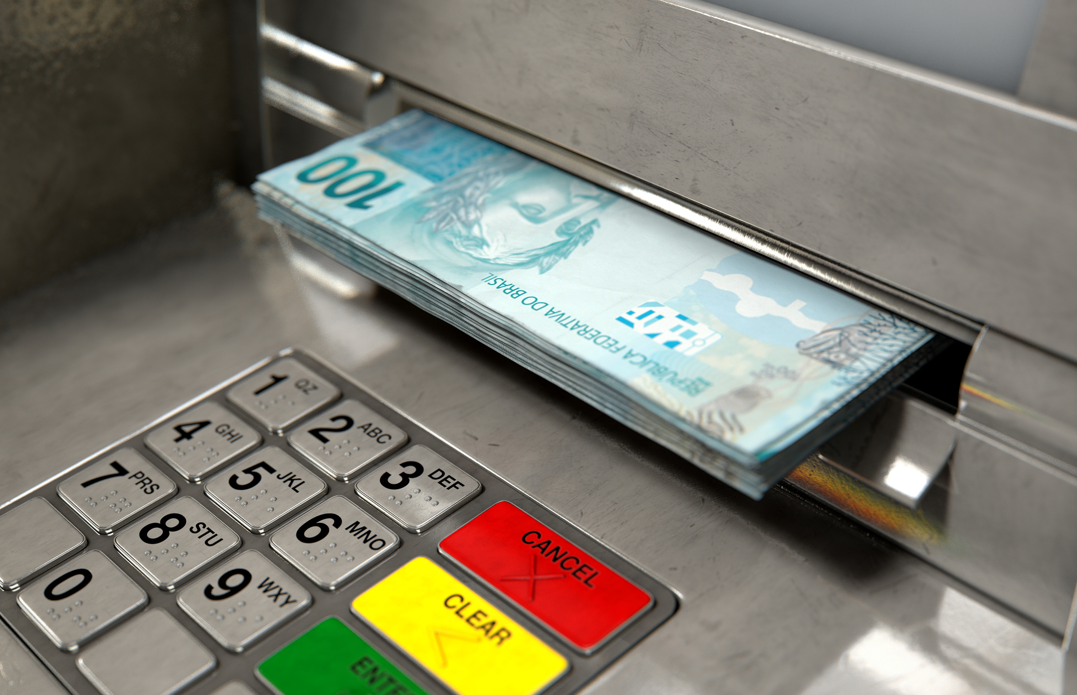 Foto horizontal mostra detalhe de teclado de caixa automático de banco, e pilha de dinheiro  com nota de 100 reais em cima saindo da máquina.