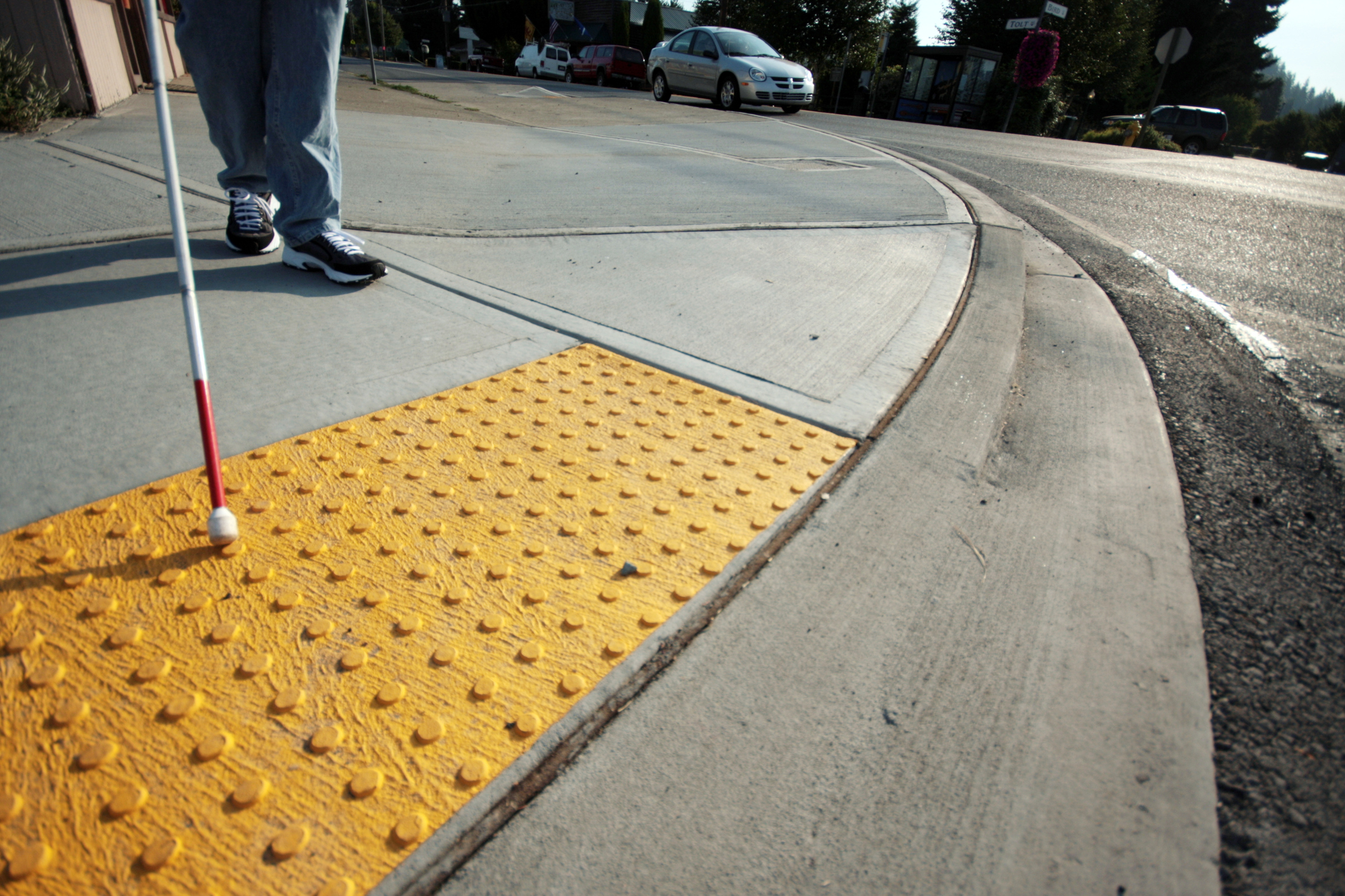 FOTO HORIZONTAL, COLORIDA, DE CALÇADA COM PISO TÁTIL AMARELO. À ESQUERDA, RECORTE DE PARTE DE PERNAS DE PESSOA CAMINHANDO COM BENGALA.