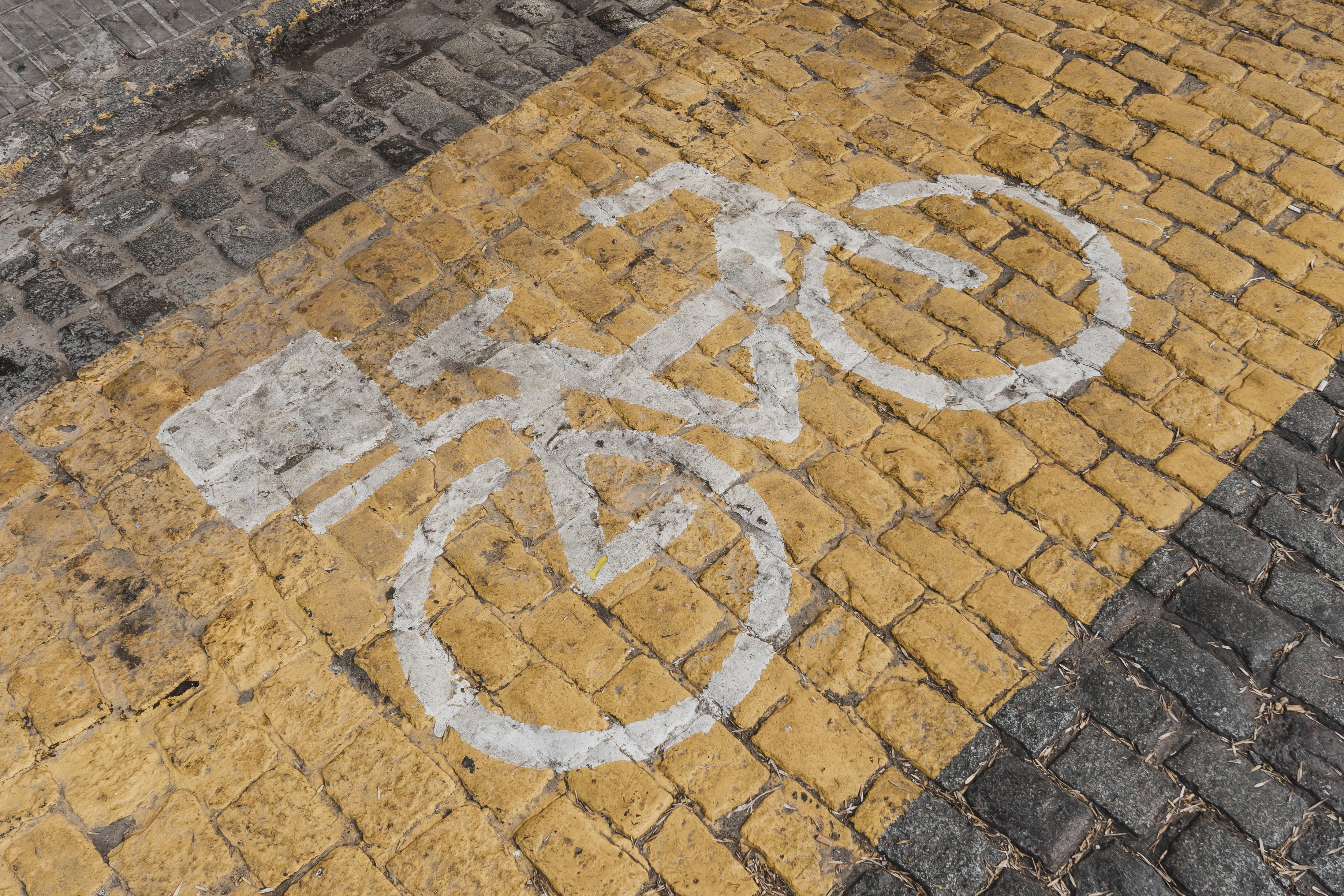 Foto horizontal, retangular, mostra uma marcação de ciclovia pintada em uma superfície de paralelepípedos. A maior parte da área visível é coberta por paralelepípedos de cor amarelada, onde o símbolo de uma bicicleta é pintado em branco. À esquerda e na parte inferior da imagem, há uma seção de paralelepípedos de cor cinza escura.

A marcação da ciclovia consiste no contorno de uma bicicleta estilizada, com as rodas, o quadro e o guidão. À esquerda da bicicleta, há uma forma retangular branca. A perspectiva da foto é de um ângulo alto, olhando para baixo na superfície da estrada, dando uma boa visão da textura dos paralelepípedos e da marcação no chão.