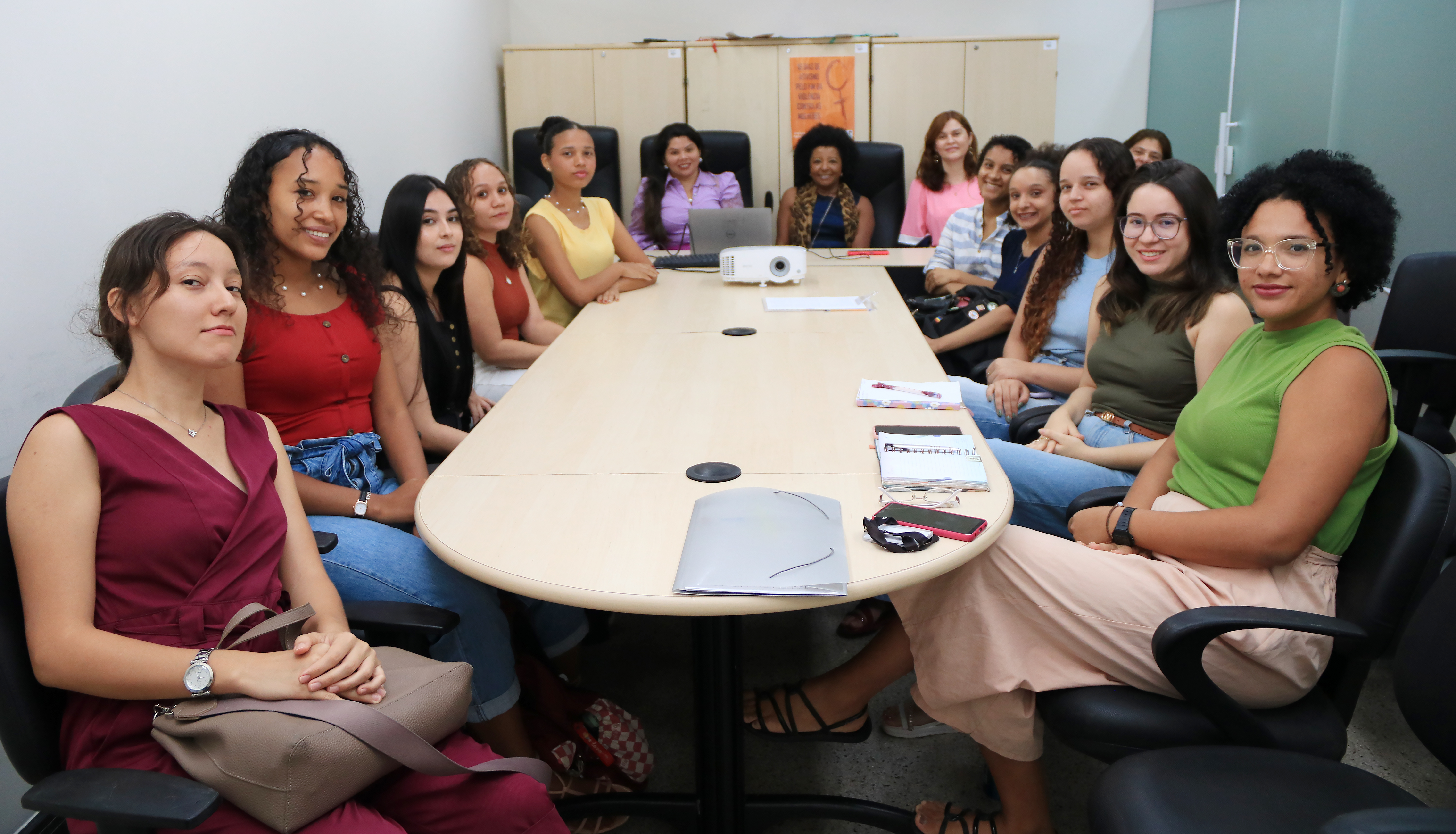 A imagem mostra um grupo de pessoas sentadas ao redor de uma mesa de conferência oval. Os rostos das pessoas estão cobertos por retângulos marrons, provavelmente por razões de privacidade. Na mesa, há vários itens, incluindo papéis, cadernos, um laptop e o que parece ser um projetor. O ambiente sugere um cenário profissional ou educacional, onde uma reunião ou aula pode estar acontecendo.