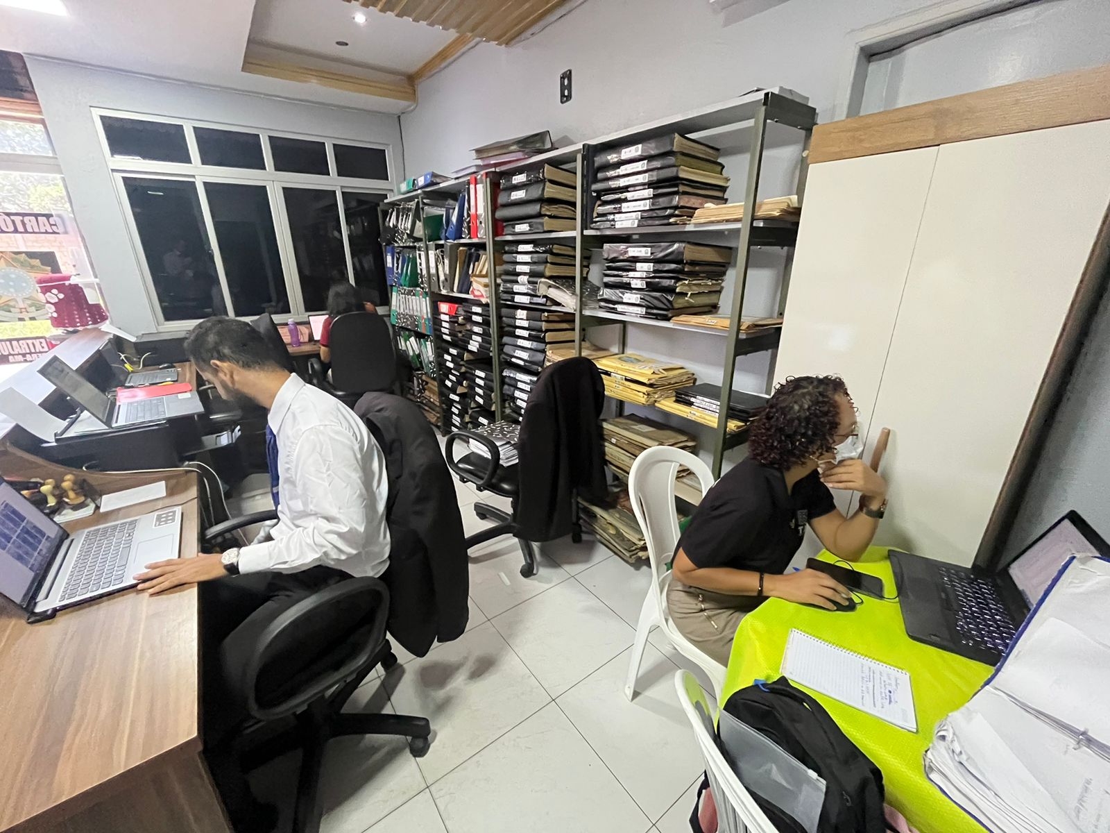 Homens e mulheres sentados diante de computador em sala dde trabalho, tendo ao fundo, estantes com documentos e processos.