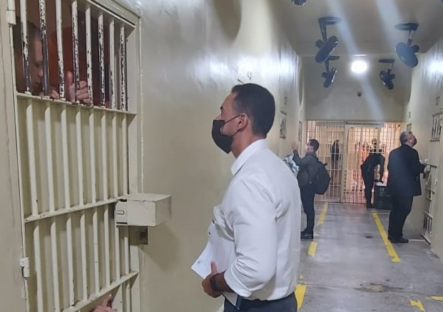 Homem pardo, de camisa social branca, conversa com pessoa presa atrás de grade em cela de presídio