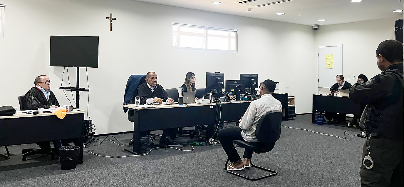 A imagem mostra uma sala de audiência judicial. No centro, há um réu sentado de costas para a câmera, vestido com uma camisa branca e calça preta. À sua direita, há um policial de pé, usando um colete à prova de balas e algemas penduradas na cintura.

Na frente do réu, do outro lado da sala, estão duas mesas com três pessoas sentadas. À esquerda, um homem usando vestes de juiz está sentado à mesa, com um computador e alguns documentos ao seu redor. Ao lado dele, outro homem, também com vestes formais, parece estar fazendo perguntas ao réu. Entre eles, uma mulher com um notebook está digitando, possivelmente uma escrivã ou assessora.

Ao fundo, na parede, há um crucifixo fixado e uma janela pequena que permite a entrada de luz natural.