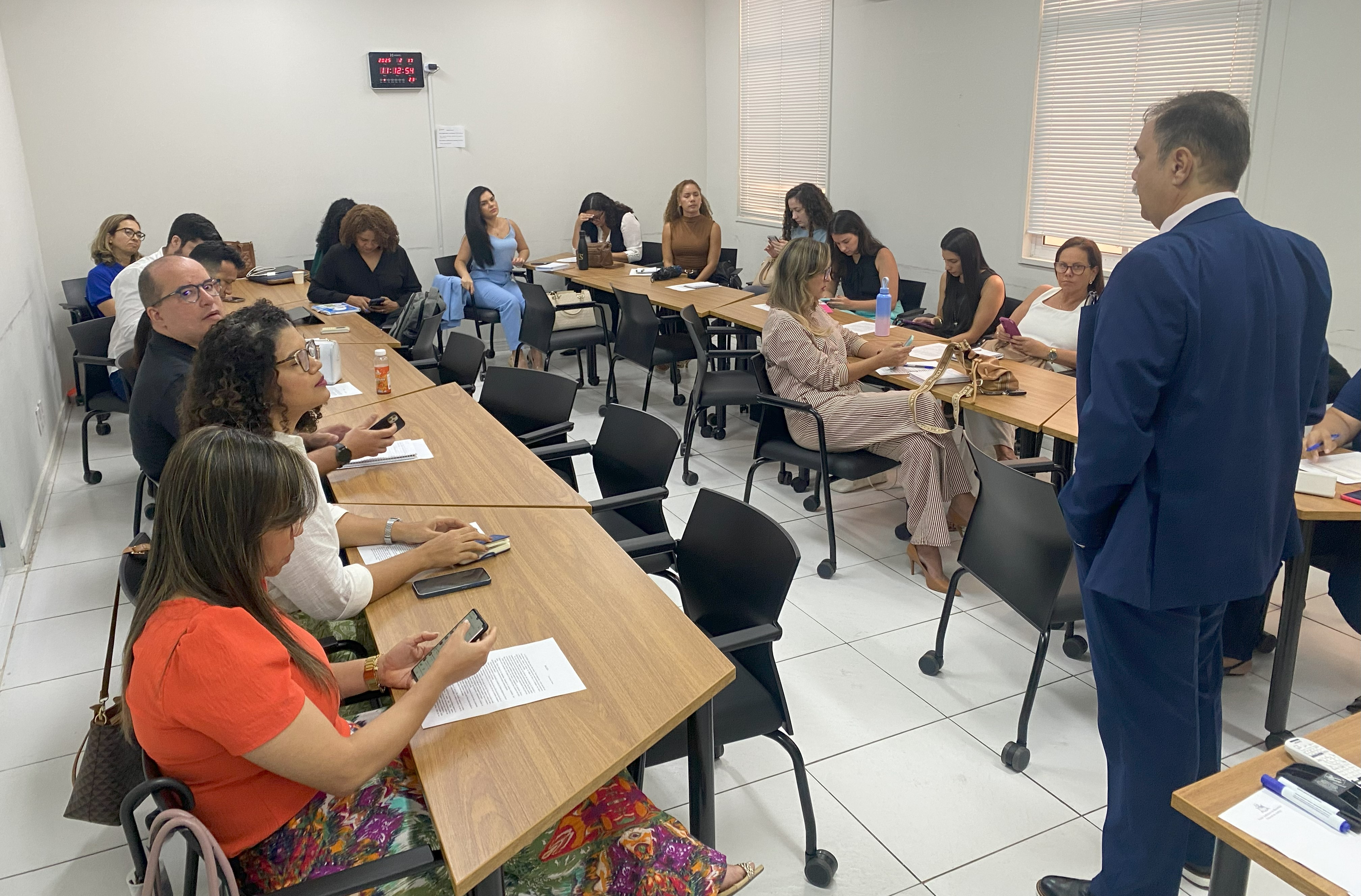 Foto horizontal, colorida, mostra uma sala de aula com paredes brancas, e um homem branco, vestido de terno azul, de pé e de costas, em primeiro plano. Ao fundo, uma bancada em formato de "U", com homens e mulheres sentados.