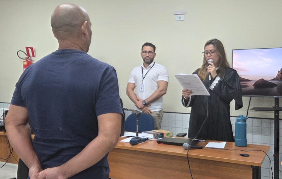 A imagem mostra uma sala de audiência judicial com três pessoas. Em primeiro plano, de costas para a câmera, está um homem calvo vestindo uma camiseta azul-escura e com as mãos para trás. À direita, atrás de uma mesa de madeira, uma mulher de óculos e cabelos longos e claros veste uma toga preta sobre uma blusa de renda branca; ela segura um papel com a mão esquerda e fala ao microfone que segura com a mão direita. No centro, ao fundo, um homem de óculos, barba e camisa branca com crachá observa a cena com os braços cruzados. Sobre a mesa de madeira há papéis, um notebook, livros jurídicos e microfones, e ao fundo, à direita, há uma televisão ligada exibindo uma paisagem.