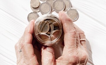 Foto horizontal, ilustrada, de mãos de pessoa idosa, branca, em torno de vidro transparente cheio de moedas. Algumas moedas estão sobre fundo branco.