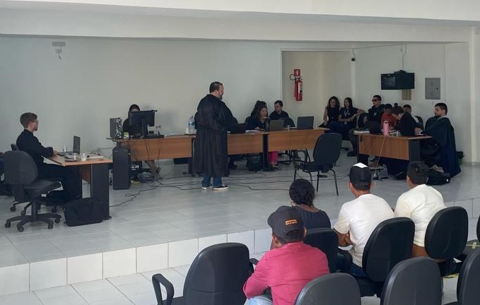 Foto horizontal, colorida, de uma sessão de tribunal, com homens e mulheres sentadas em mesas e cadeiras. Em primeiro plano, uma fileira de cadeiras está disposta para os espectadores, com algumas pessoas sentadas. Ao fundo,: mesas estão alinhadas, com pessoas sentadas e  algumas usando laptops.O salão tem paredes brancas, e há uma porta visível no lado direito da foto.  Algumas pessoas estão vestindo vestes pretas As pessoas sentadas nas cadeiras no primeiro plano assistem à sessão.