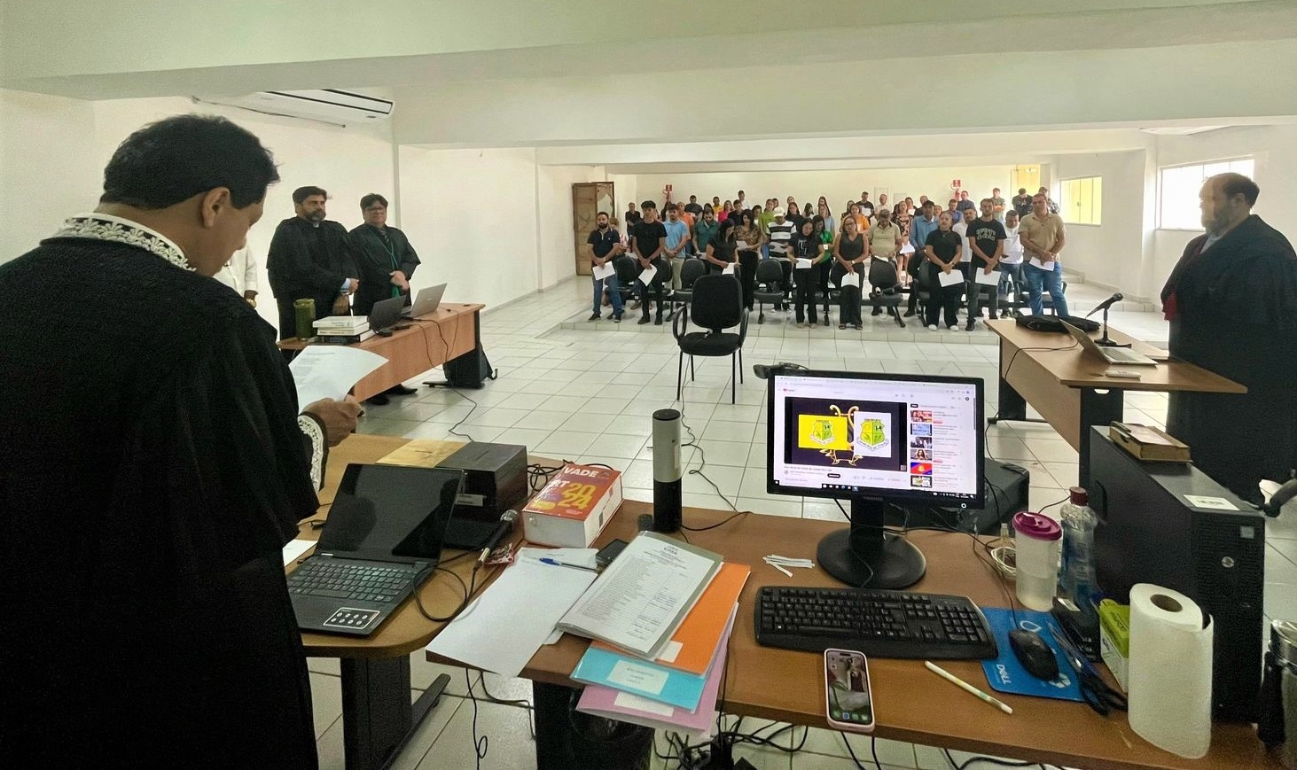 imagem na qual aparece um homem de preto, de costas, lendo um documento, em uma sala com pessoas na plateia assistindo, durante uma sessão de julgamento