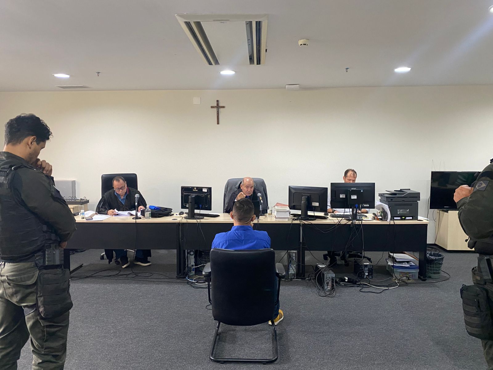 A imagem mostra uma cena de um tribunal. No centro, há um réu sentado em uma cadeira de costas para a câmera, vestindo uma camisa azul, sendo interrogado por dois juízes que estão sentados atrás de uma mesa comprida. Na frente deles, há monitores de computador, papéis e outros itens de escritório. Dois agentes de segurança estão de pé nas laterais da sala, observando o procedimento. A sala é simples, com paredes claras e uma cruz afixada acima dos juízes, sugerindo que se trata de um tribunal brasileiro, onde esse símbolo costuma ser encontrado em salas de audiência.