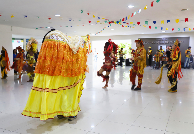 Apresentação do Bumba Meu Boi em um salão. Vários brincantes dançam, com destaque para o boi amarelo no centro. O teto está decorado com bandeirinhas coloridas.