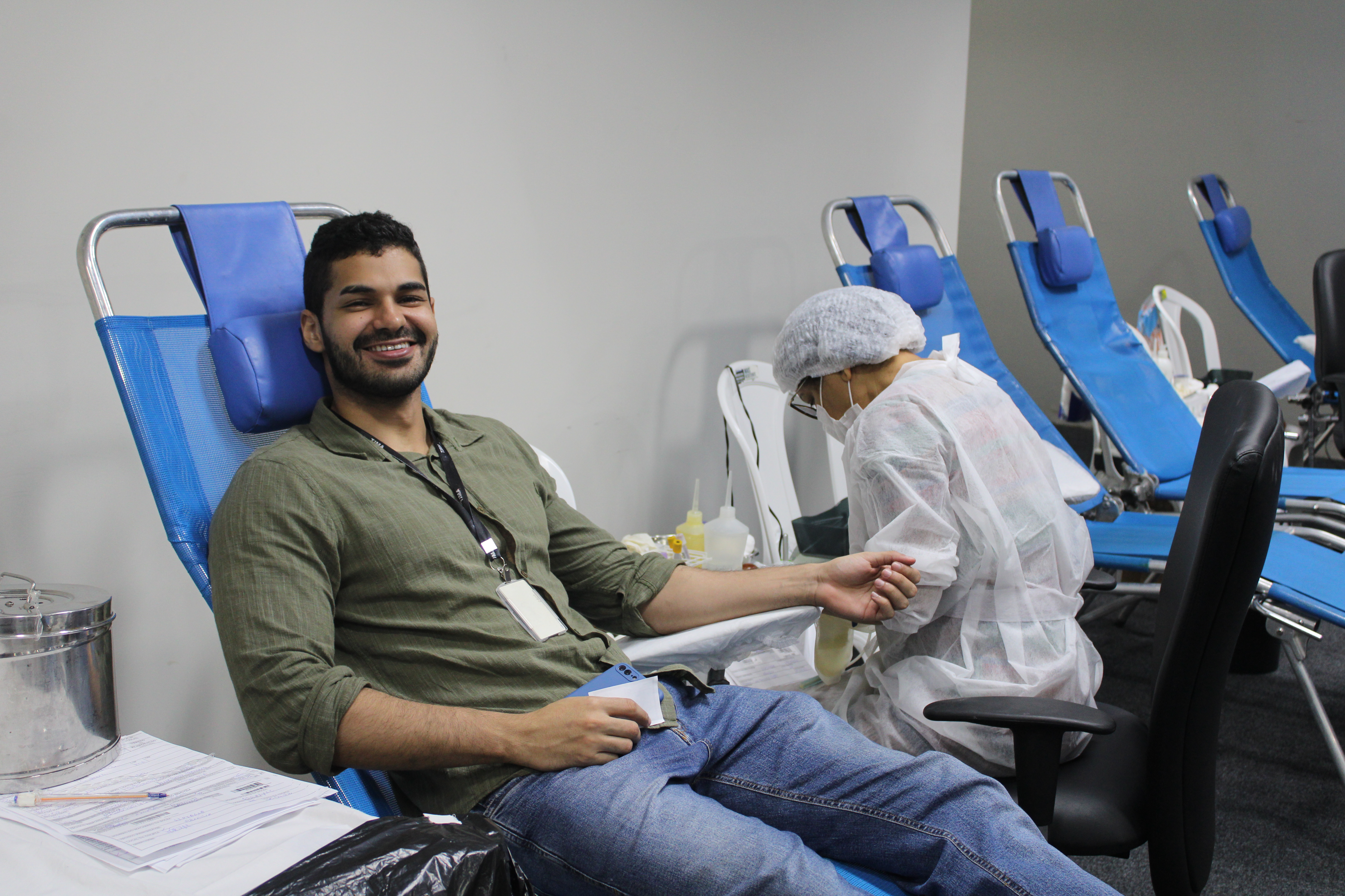 Foto colorida. Servidor sorri deitado em cadeira azul reclinável enquanto doa sangue.
