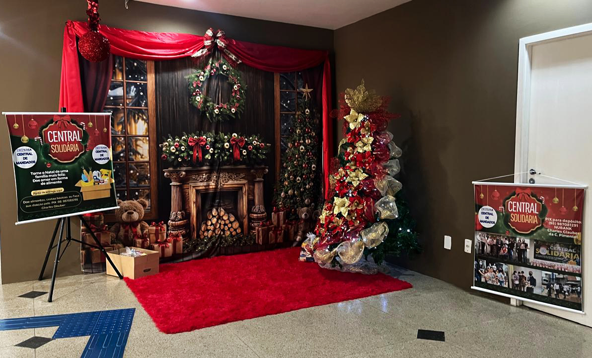 A imagem mostra um estande decorado para uma campanha de doações chamada "Central Solidária", aparentemente em clima natalino. A decoração inclui uma árvore de Natal com laços vermelhos e dourados e enfeites brilhantes, além de um cenário com uma lareira e guirlandas, recriando um ambiente aconchegante. A árvore está cercada por sacos de presentes e uma pelúcia de urso. Há uma placa vertical em frente ao estande, promovendo a campanha com informações sobre doação de alimentos e brinquedos.

O estande está montado em um espaço interno, com piso de granito ou mármore e um tapete vermelho que complementa o tema festivo. O fundo simula uma sala de estar com janelas decoradas para o Natal, coroas de flores, e uma lareira cheia de lenha, adicionando um toque acolhedor ao cenário.