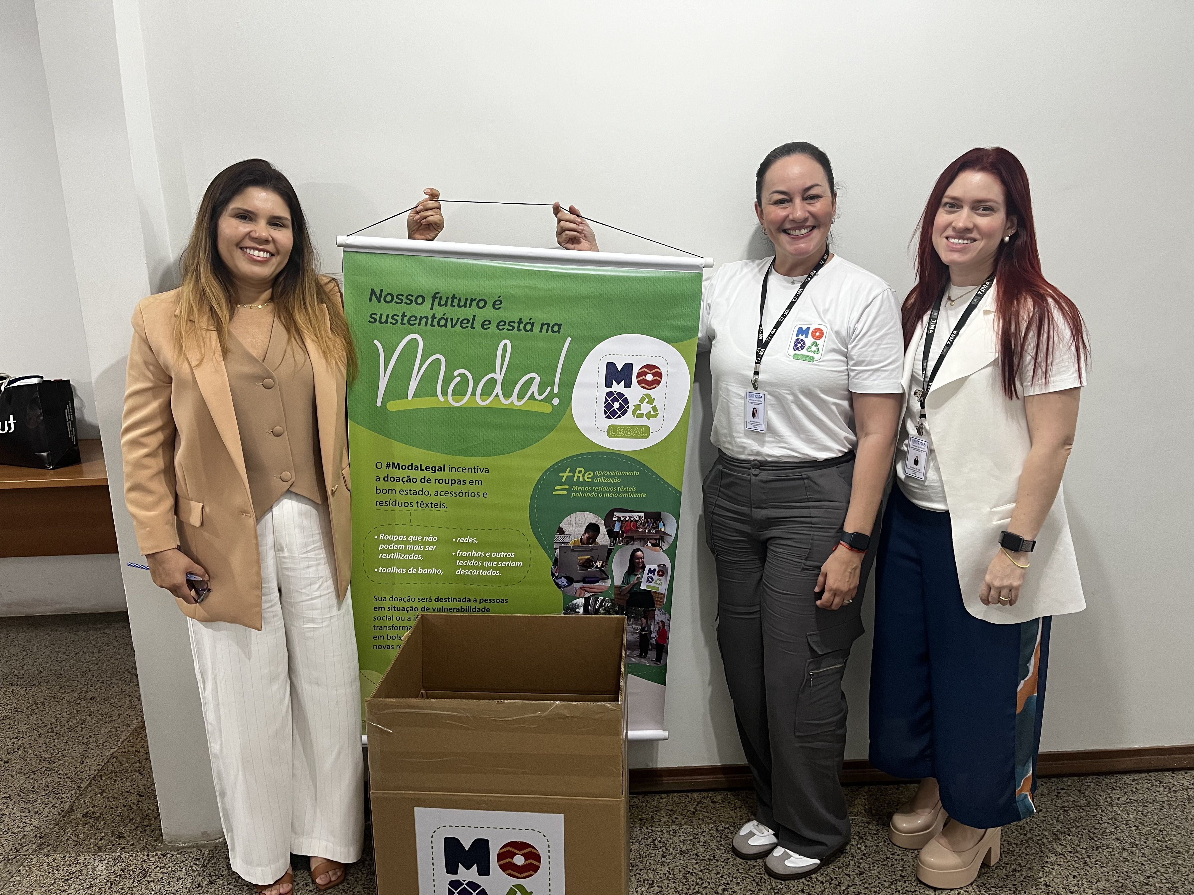 Três mulheres estão em pé ao lado de um banner do projeto Moda Legal. O banner contém informações sobre a iniciativa, que incentiva a doação de roupas, acessórios e resíduos têxteis. Duas mulheres vestem camisetas brancas com o logotipo do projeto, e a terceira usa um blazer bege. À frente delas, há uma caixa coletora identificada com o logotipo do projeto, destinada ao recebimento de doações. O ambiente tem paredes brancas e piso de granito.