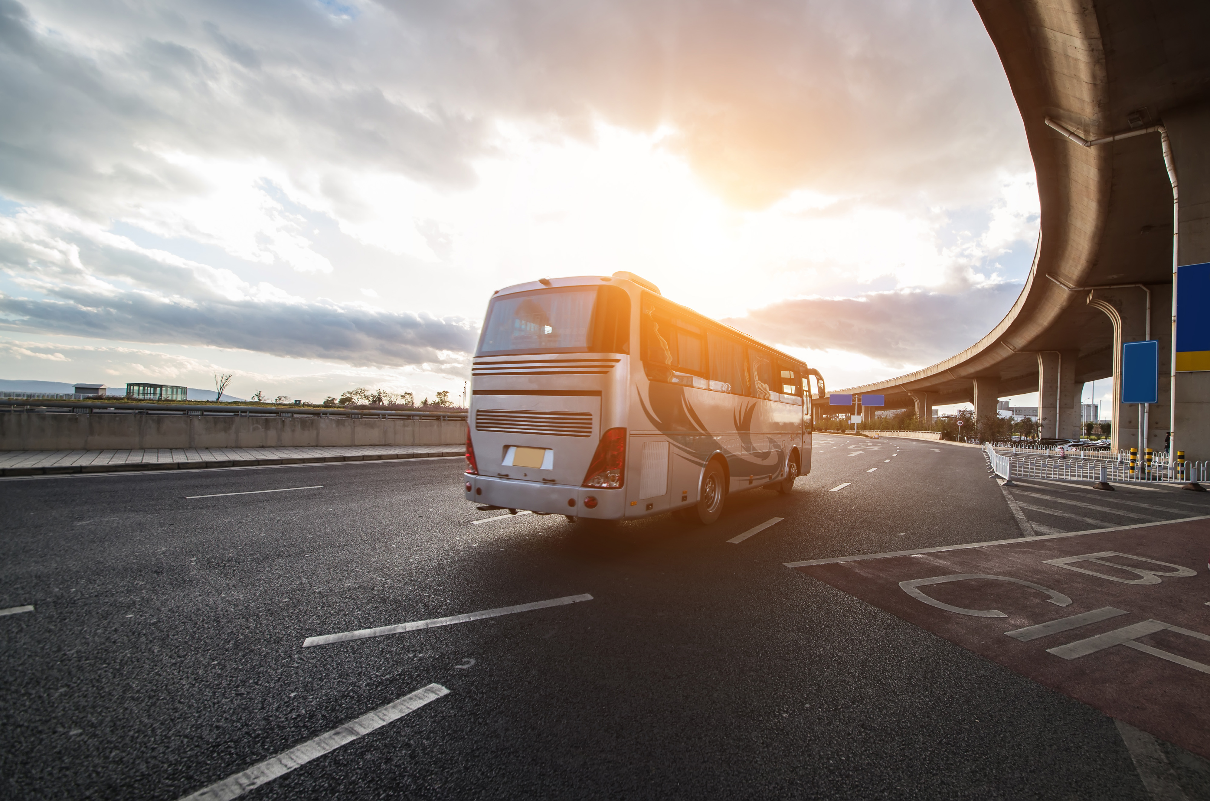 Foto horizontal, colorida, mostra  um ônibus prateado em pista de asfalto sob um viaduto de concreto curvo. O ônibus está na faixa da direita, afastando-se do observador. A luz do sol incide na lateral do ônibus, criando um brilho quente.

A estrada tem várias faixas de rodagem marcadas com linhas brancas. À esquerda da estrada, há uma barreira de concreto e, além dela, uma paisagem distante com montanhas sob um céu nublado com raios de sol rompendo as nuvens. À direita da estrada, sob o viaduto, há sinalizações de trânsito e o início de uma área com marcações no asfalto. A atmosfera geral da foto sugere o final da tarde ou o início da manhã devido à luz suave e ao ângulo do sol.