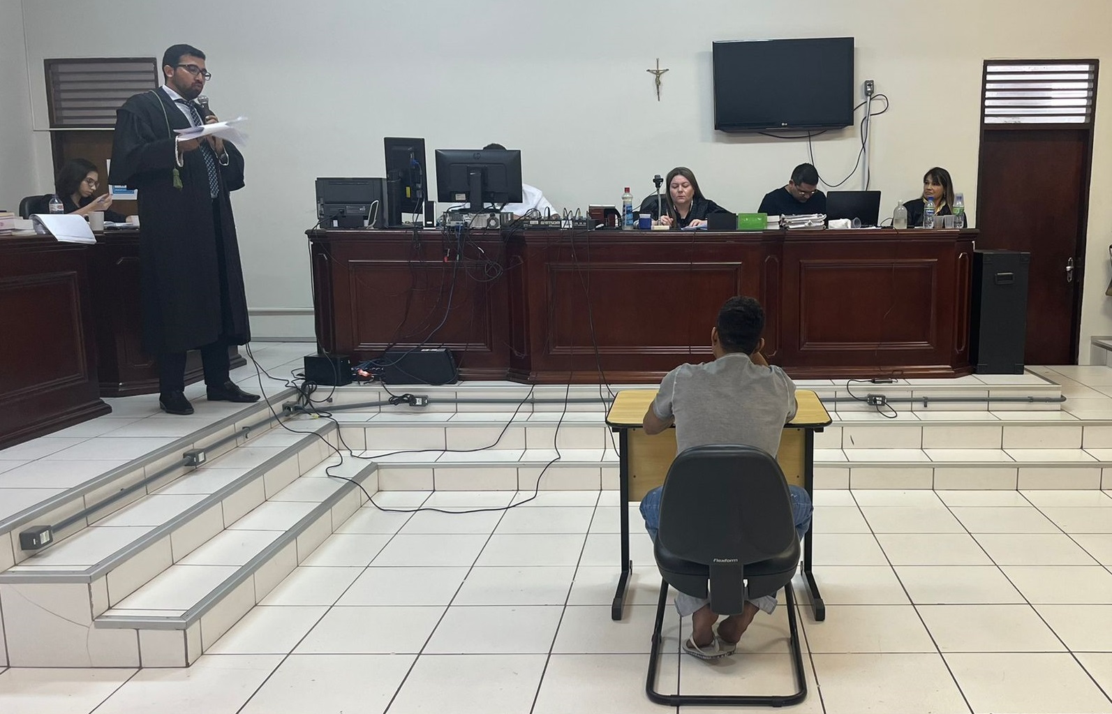 Homem pardo, réu, sentado de costas no salão do Tribunal do Júri, com promotor de pé ao seu lado e juíza sentada