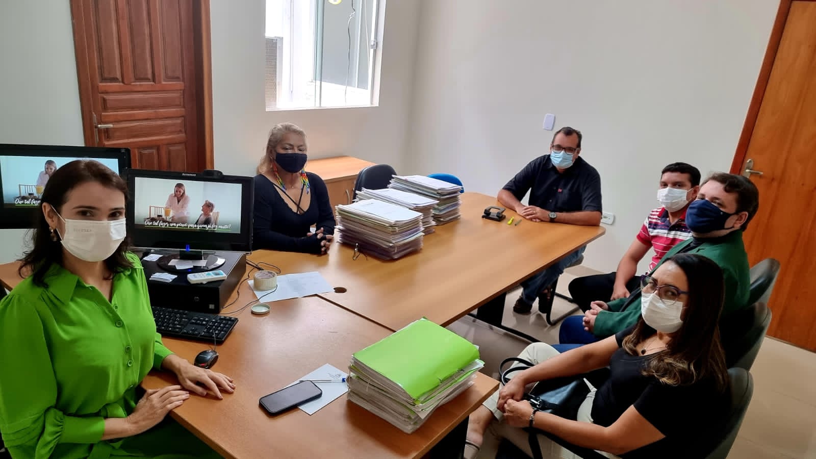 Sala de reunião com mulher branca, vestida de verde, com homens e mulheres sentados à mesa de trabalho.